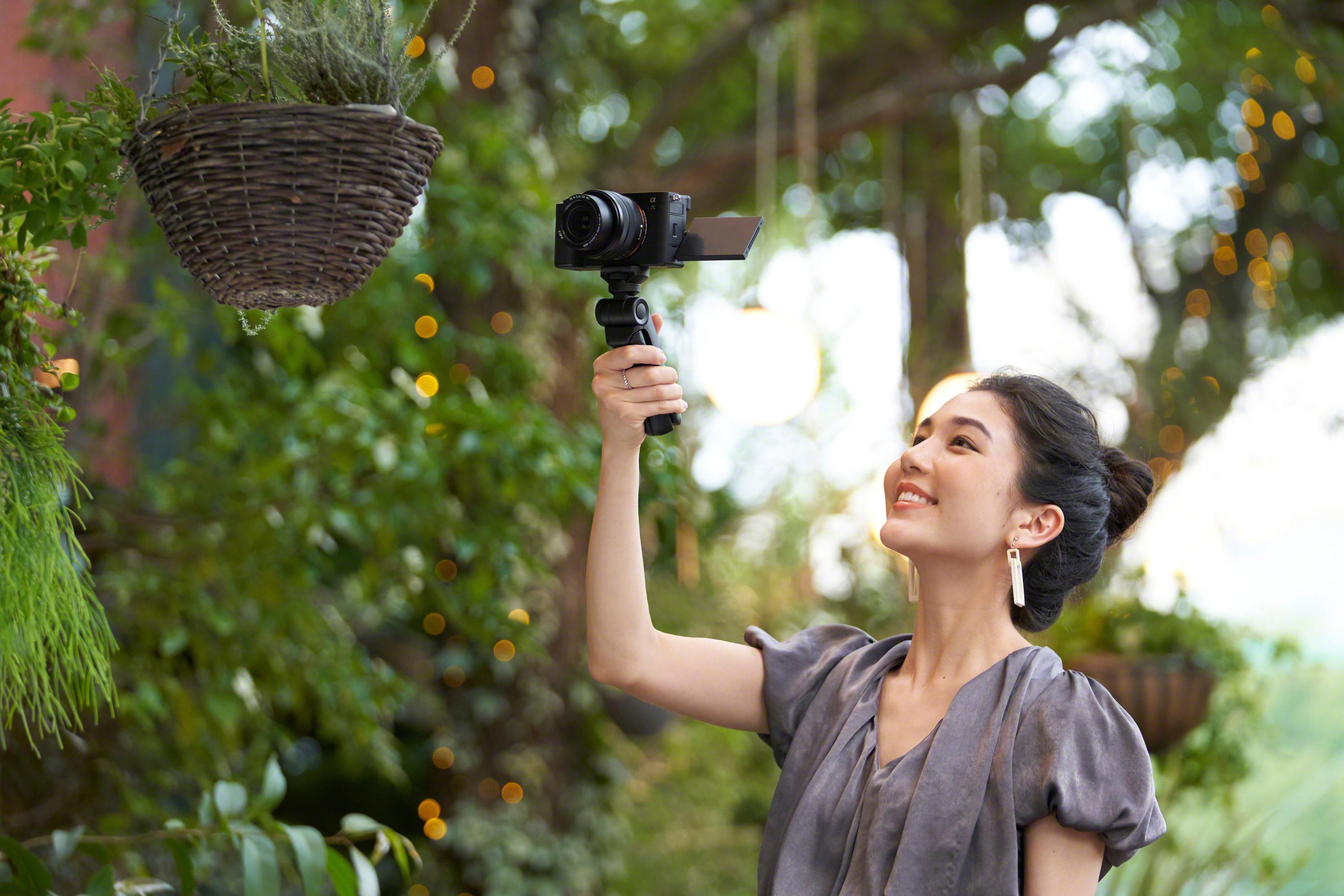 Een vrouw die een camera vasthoudt met een open scherm, glimlachend. Planten en lichten achtergrond.