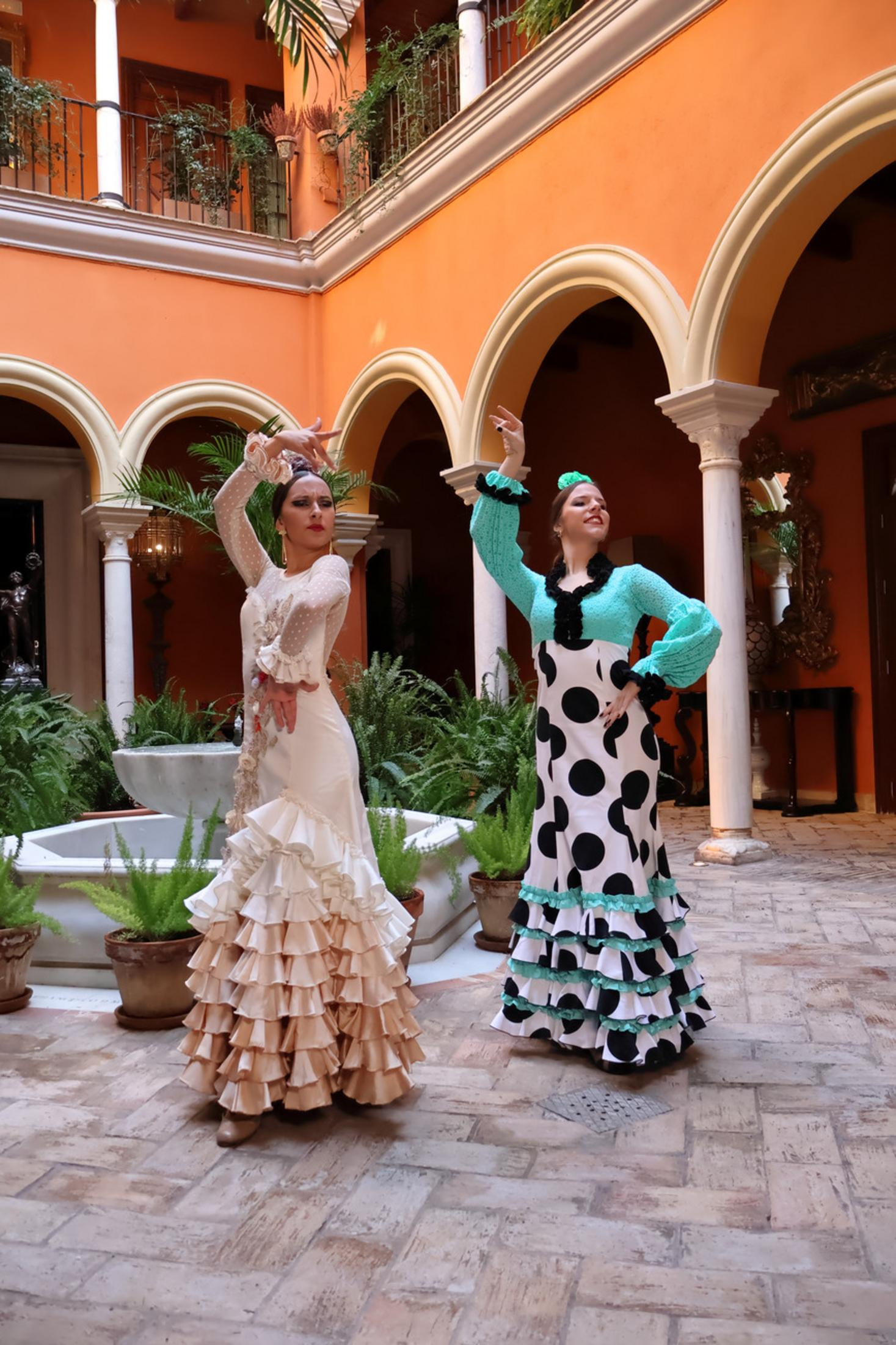 Dwie kobiety w kolorowych sukniach flamenco pozują na dziedzińcu z łukami i pomarańczowymi ścianami.