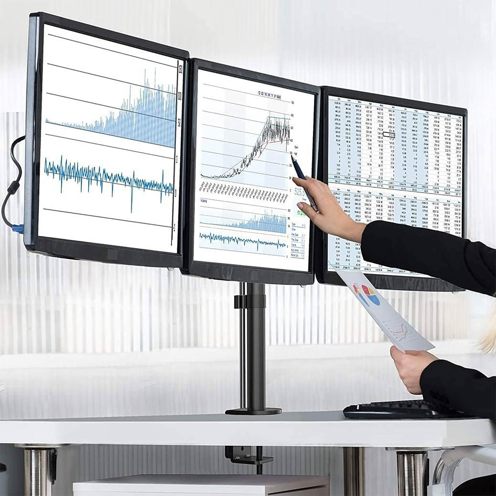 Tre monitor per computer su una scrivania. Una mano indica lo schermo centrale, tenendo un foglio.