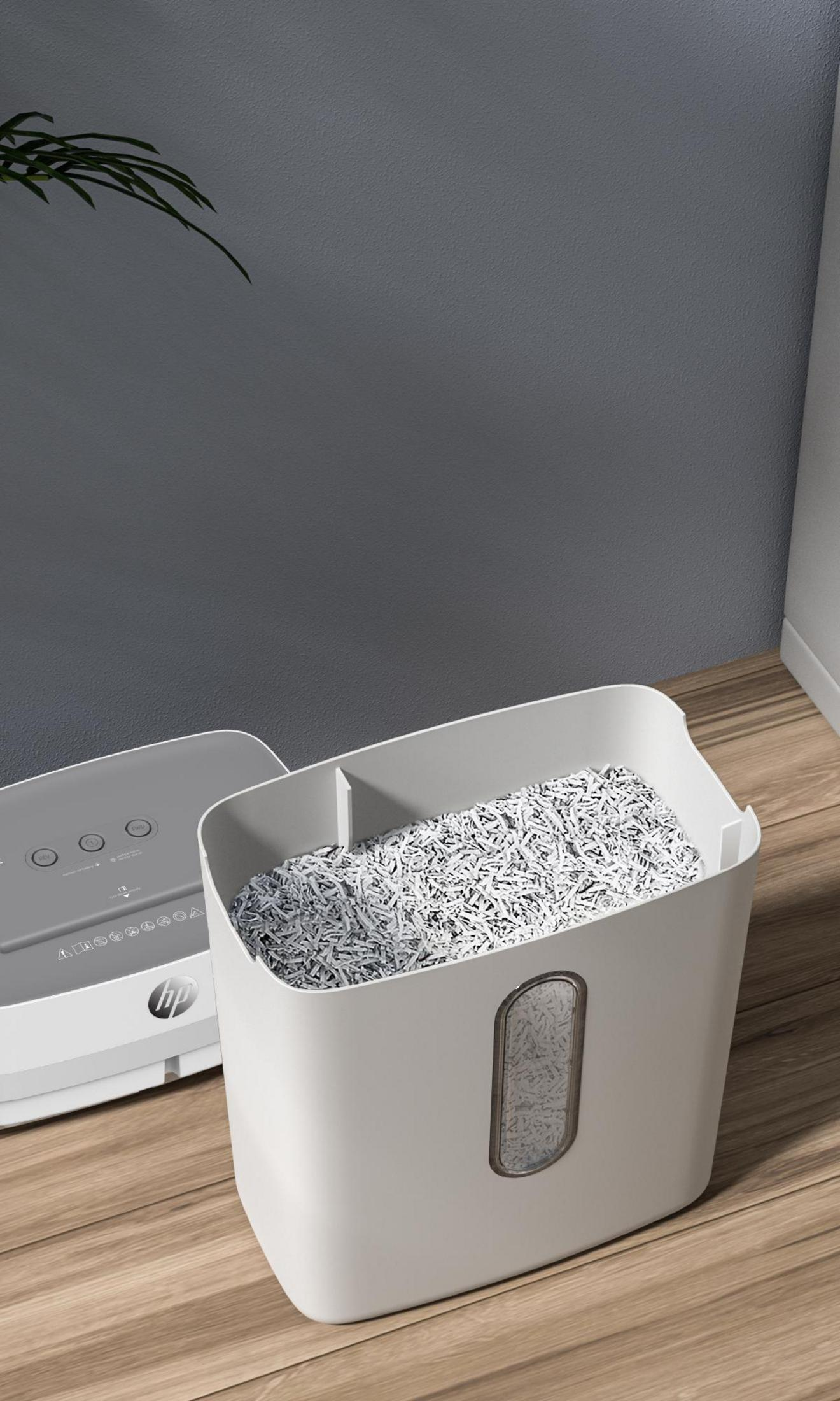 Biała niszczarka do papieru z białym pojemnikiem. Pocięty papier wypełnia pojemnik. Szara ściana i drewniana podłoga.