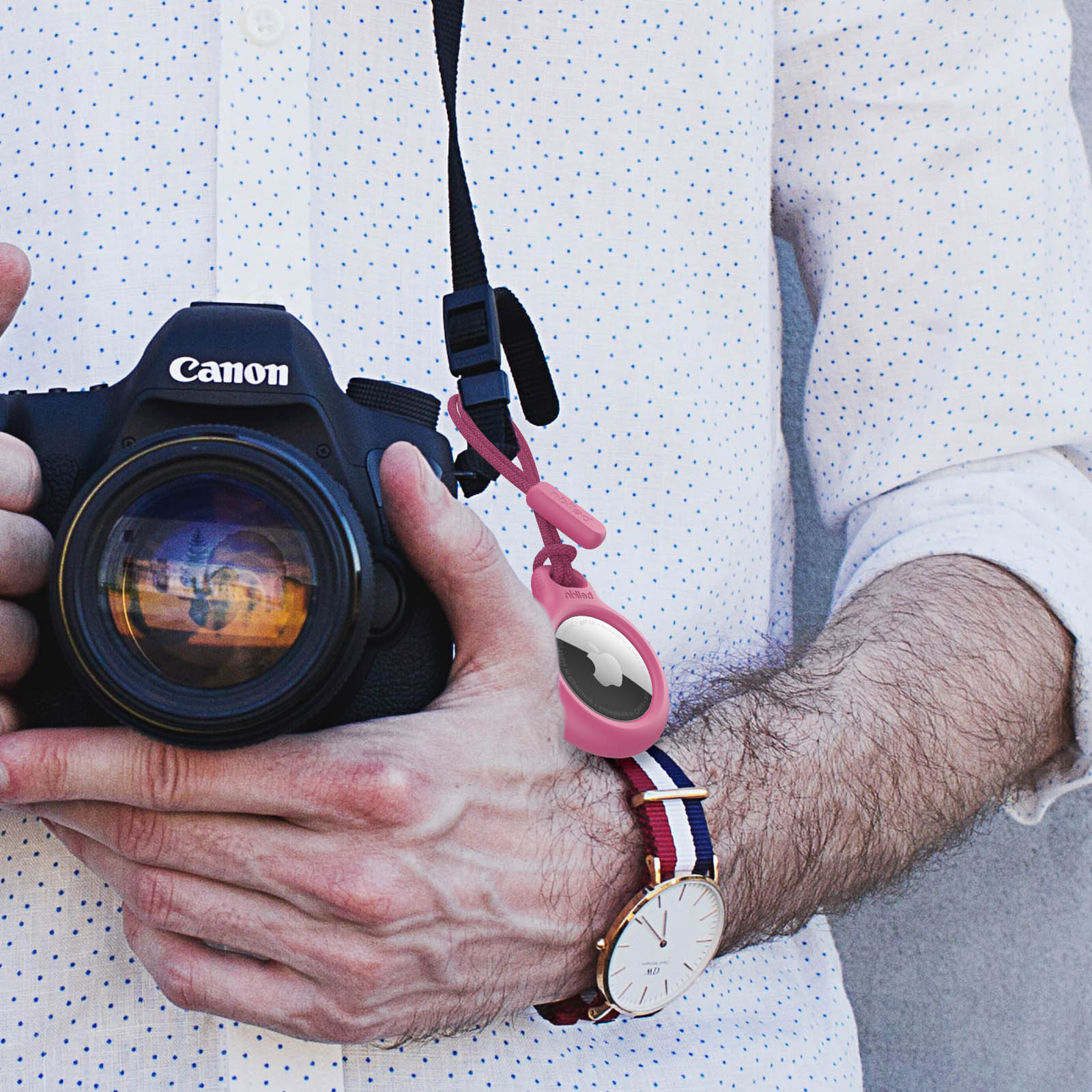 Osoba trzymająca aparat Canon z zegarkiem i różowym AirTagiem przymocowanym do paska.