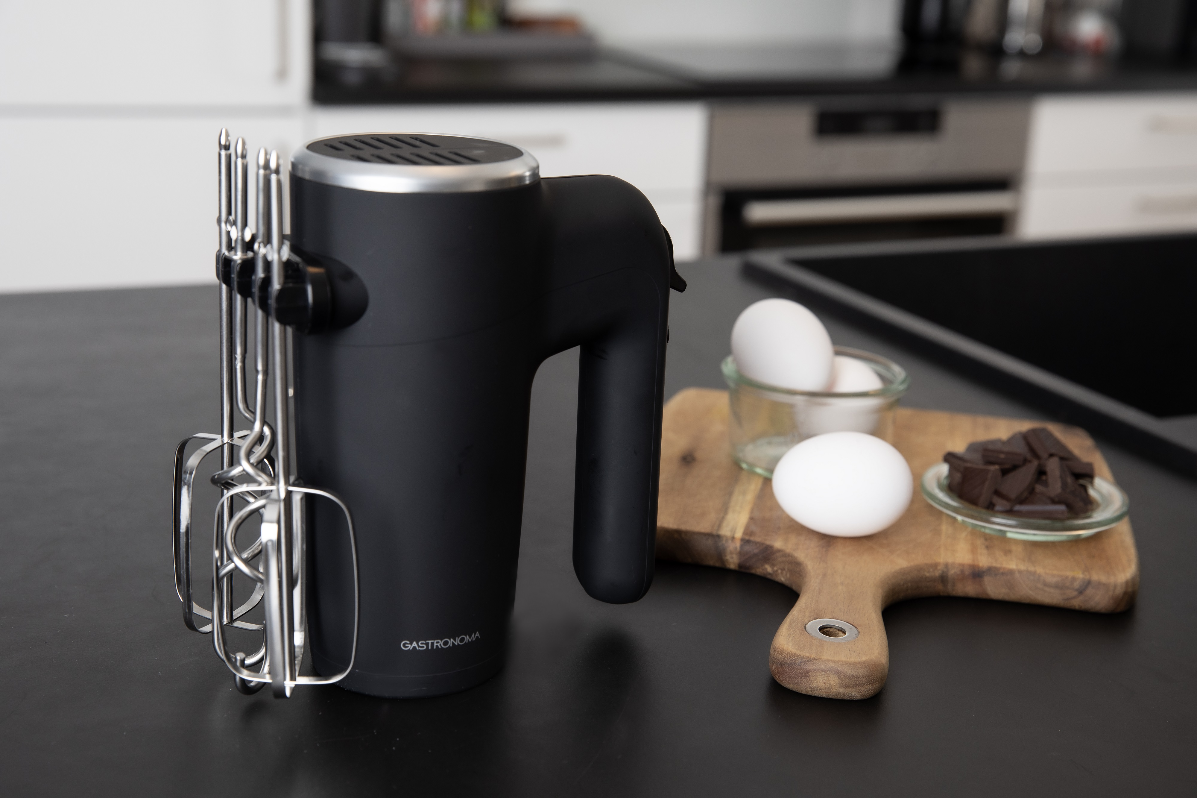 Czarny mikser ręczny z przystawkami obok jajek i czekolady na drewnianej desce.
