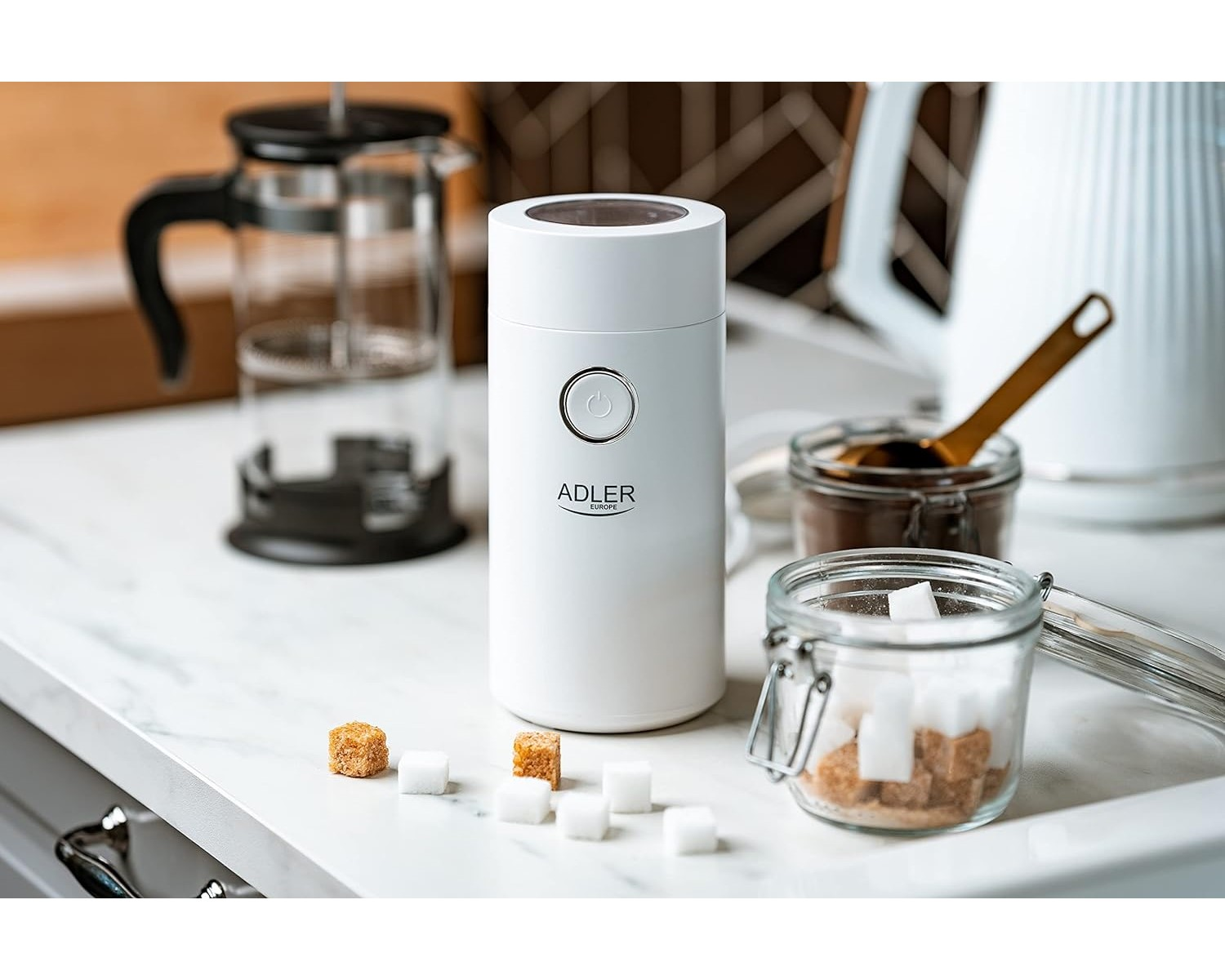 Eine weiße Kaffeemühle, Zuckerwürfel, Kaffeepulver und eine French Press stehen auf einer weißen Oberfläche.