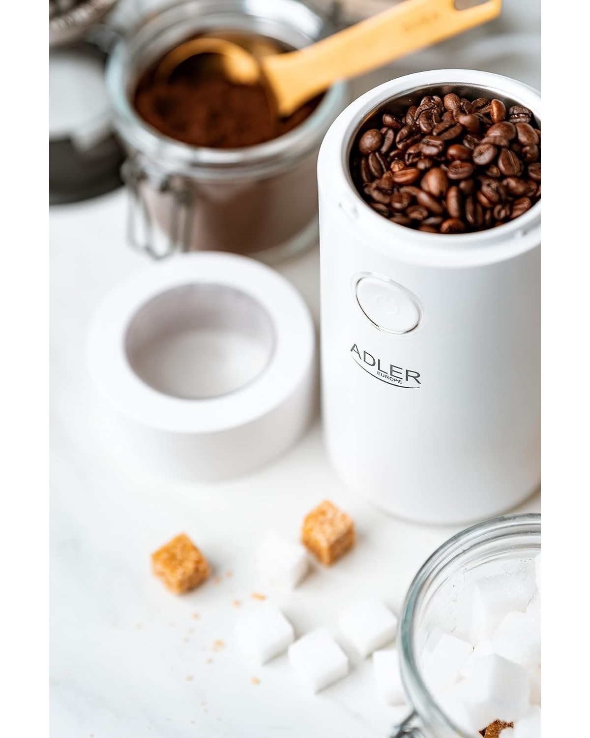 Weiße Kaffeemühle mit Kaffeebohnen. Zuckerwürfel und gemahlener Kaffee in Gläsern auf weißer Oberfläche.