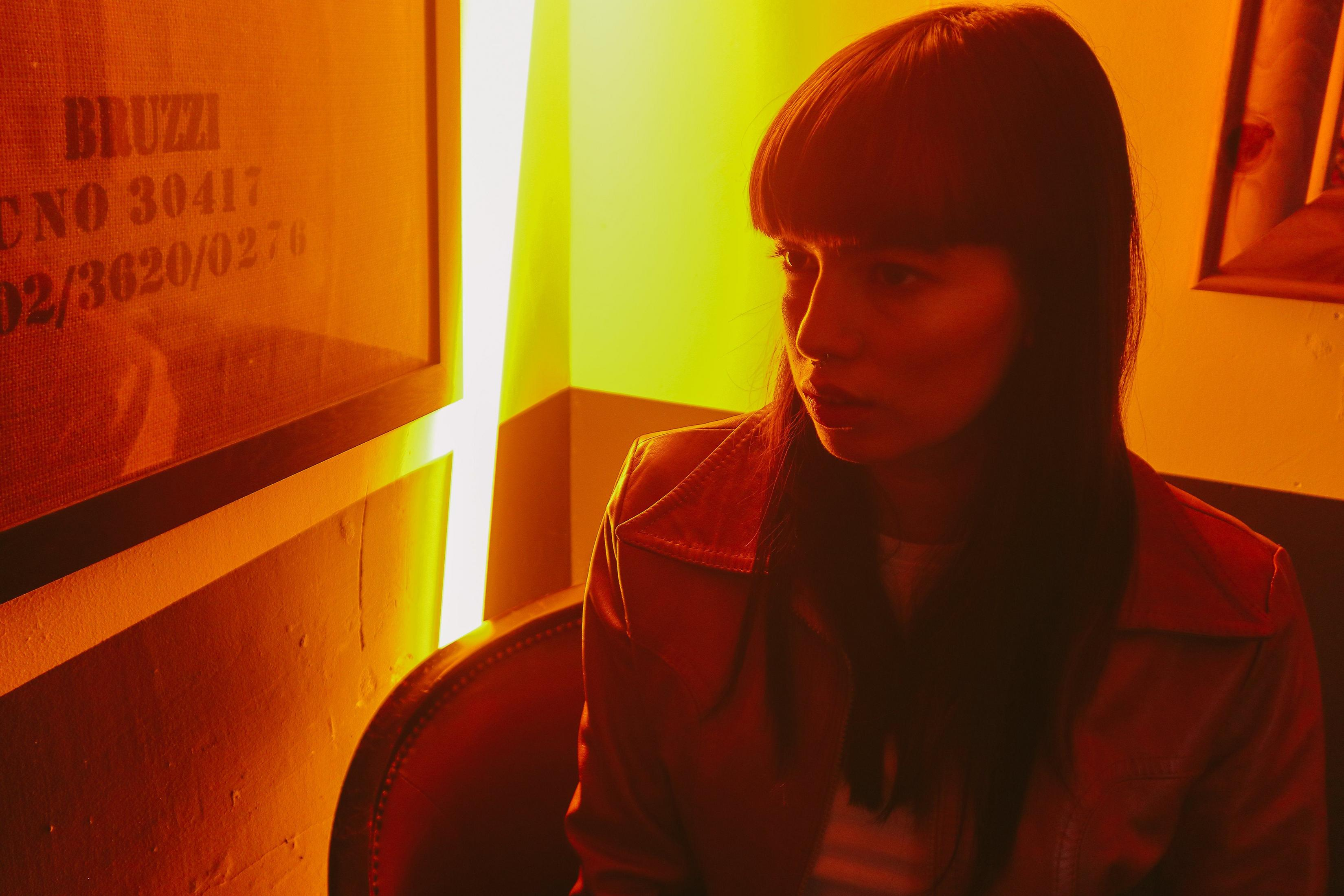 Eine Frau in einer roten Jacke in einem Raum mit Neonlicht. Der Raum hat ein Poster und ist schwach beleuchtet.
