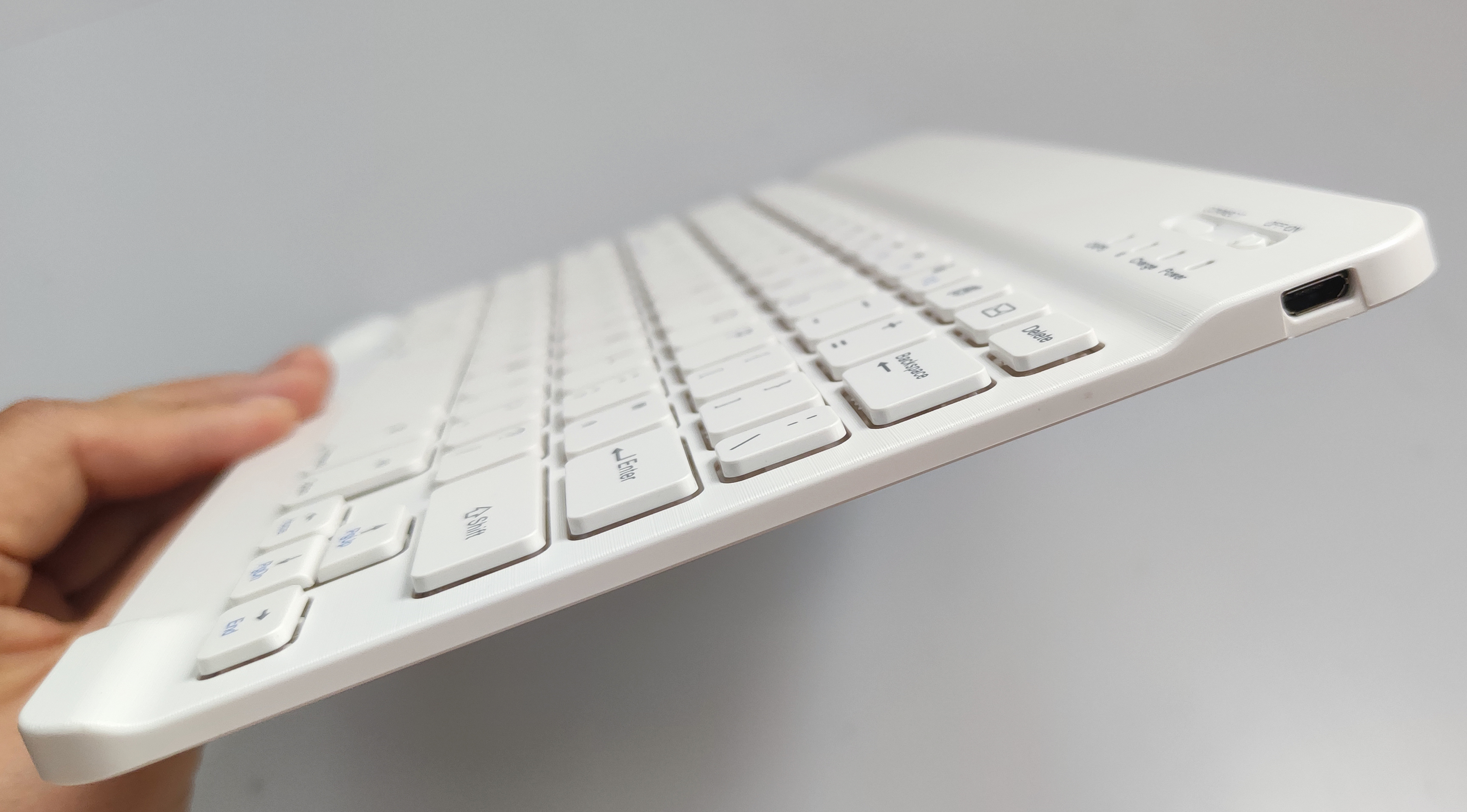 Weiße kabellose Tastatur, die von einer Hand gehalten wird. Zeigt Tasten und einen USB-C-Anschluss.