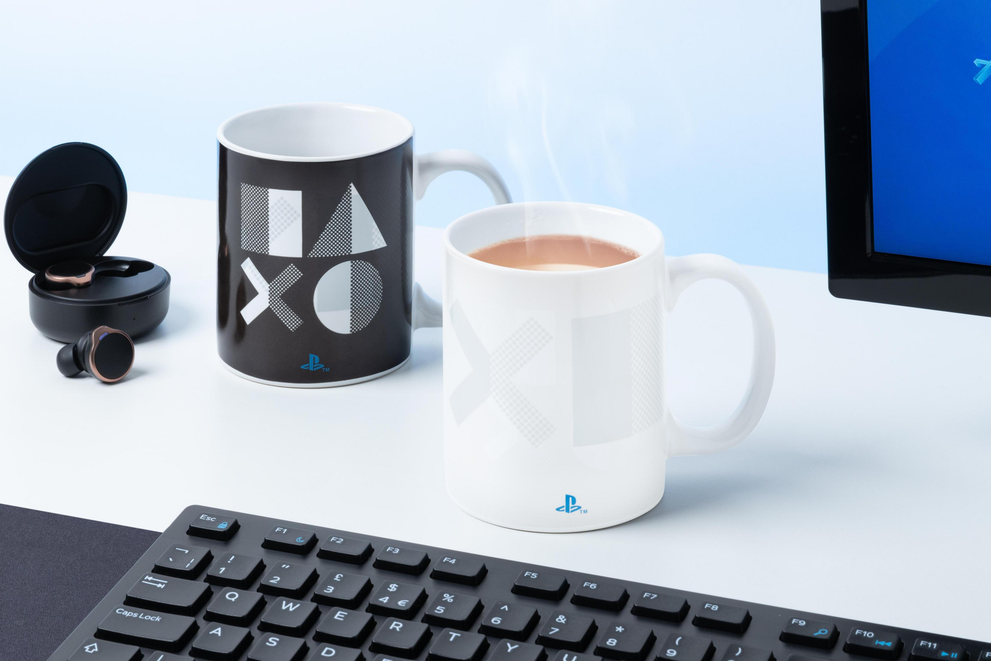 Zwei PlayStation-Tassen, eine schwarz mit Symbolen, eine weiß mit Symbol. Ohrhörer, Tastatur und Monitor sind sichtbar.