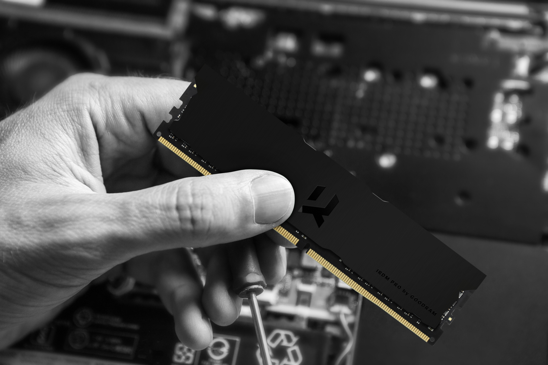 Ręka trzyma czarny moduł RAM ze złotymi złączami i śrubokręt, na tle płyty głównej komputera. Czarno-białe zdjęcie.