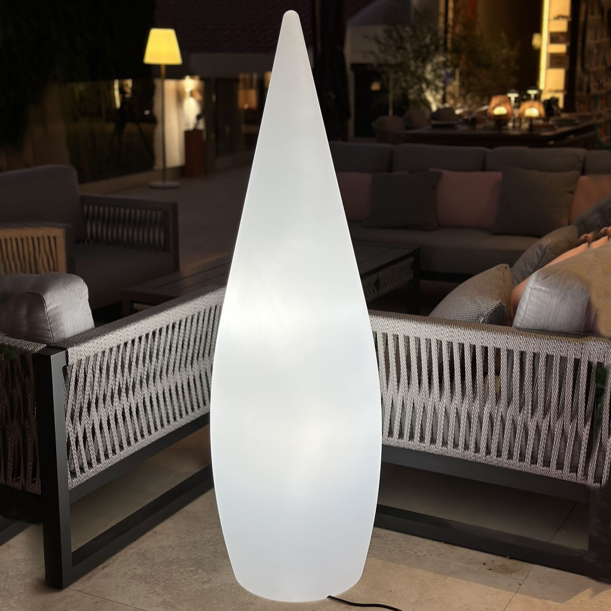 Eine hohe, weiße, leuchtende Außen-Stehleuchte. Tropfenförmig auf einer Terrasse stehend.
