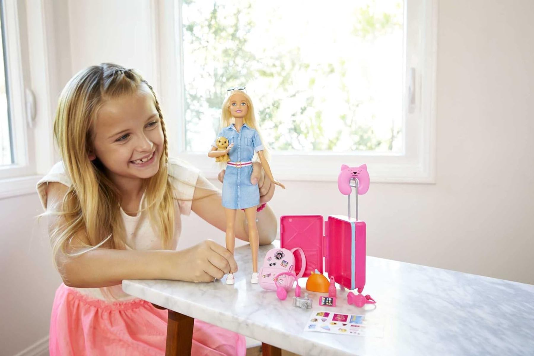 Una ragazza sorride a una bambola Barbie con una valigia, uno zaino e accessori su un tavolo vicino a una finestra.