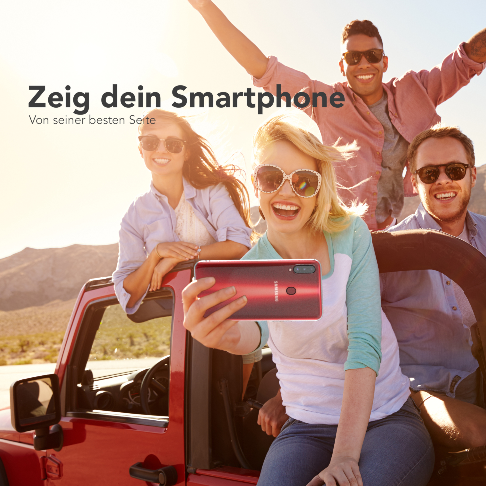 Gruppe von Leuten macht ein Selfie mit einem roten Telefon in sonniger Umgebung.