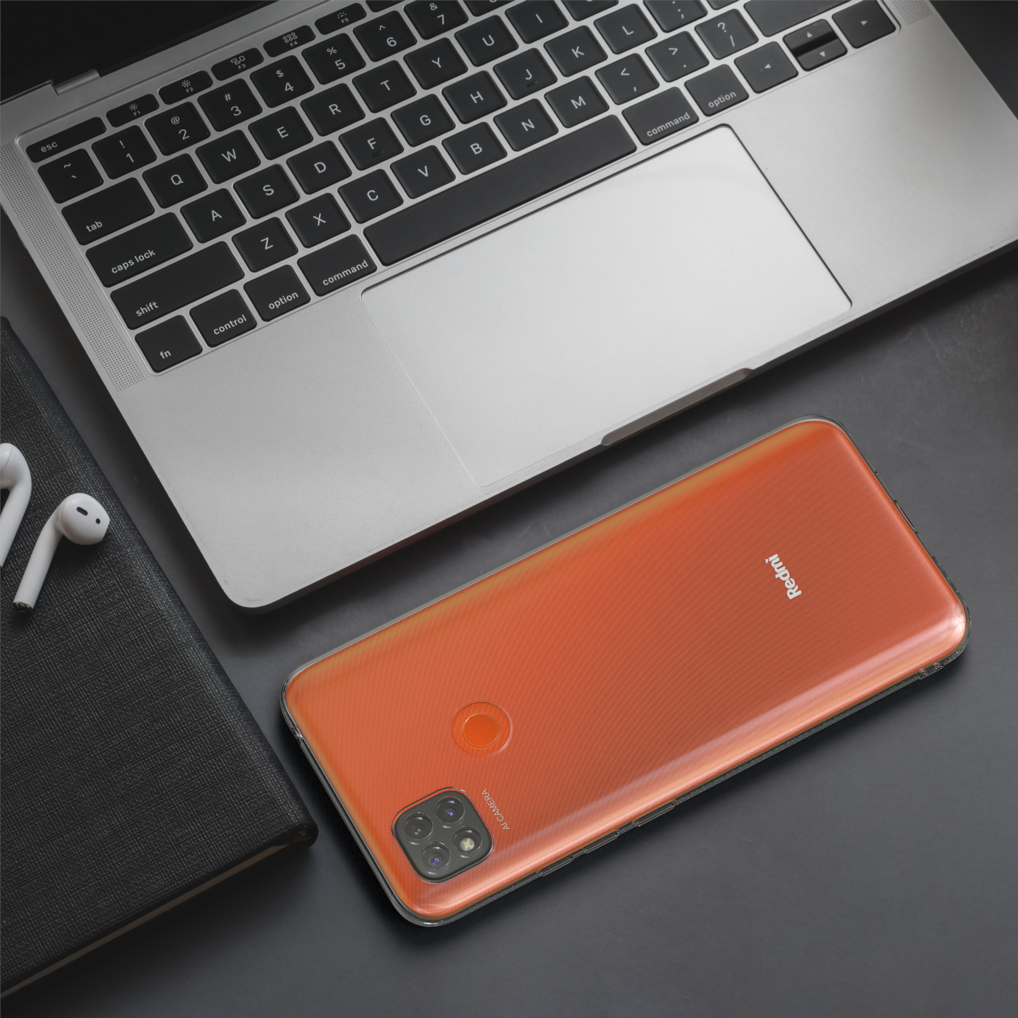 Orangefarbenes Telefon mit Kamera und ein Notizbuch auf einem Tisch mit einem Laptop. Weiße Ohrhörer vorhanden.