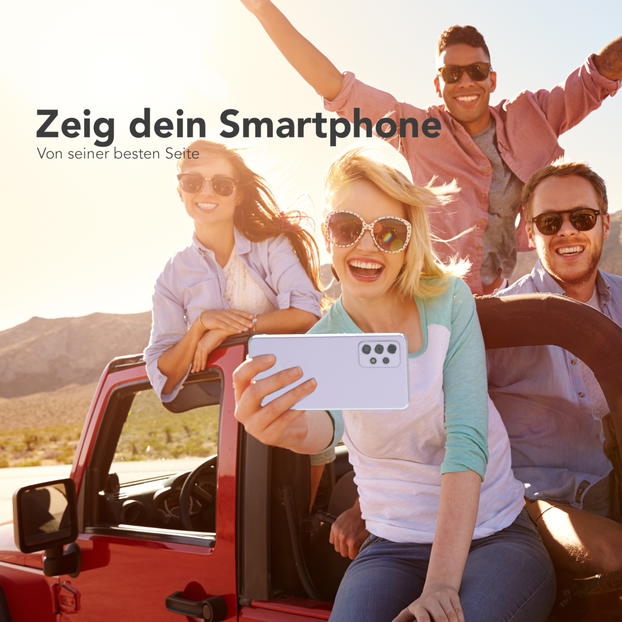 Vier lächelnde Personen in einem roten Jeep machen ein Selfie mit einem Telefon. Der Hintergrund ist sonnig.