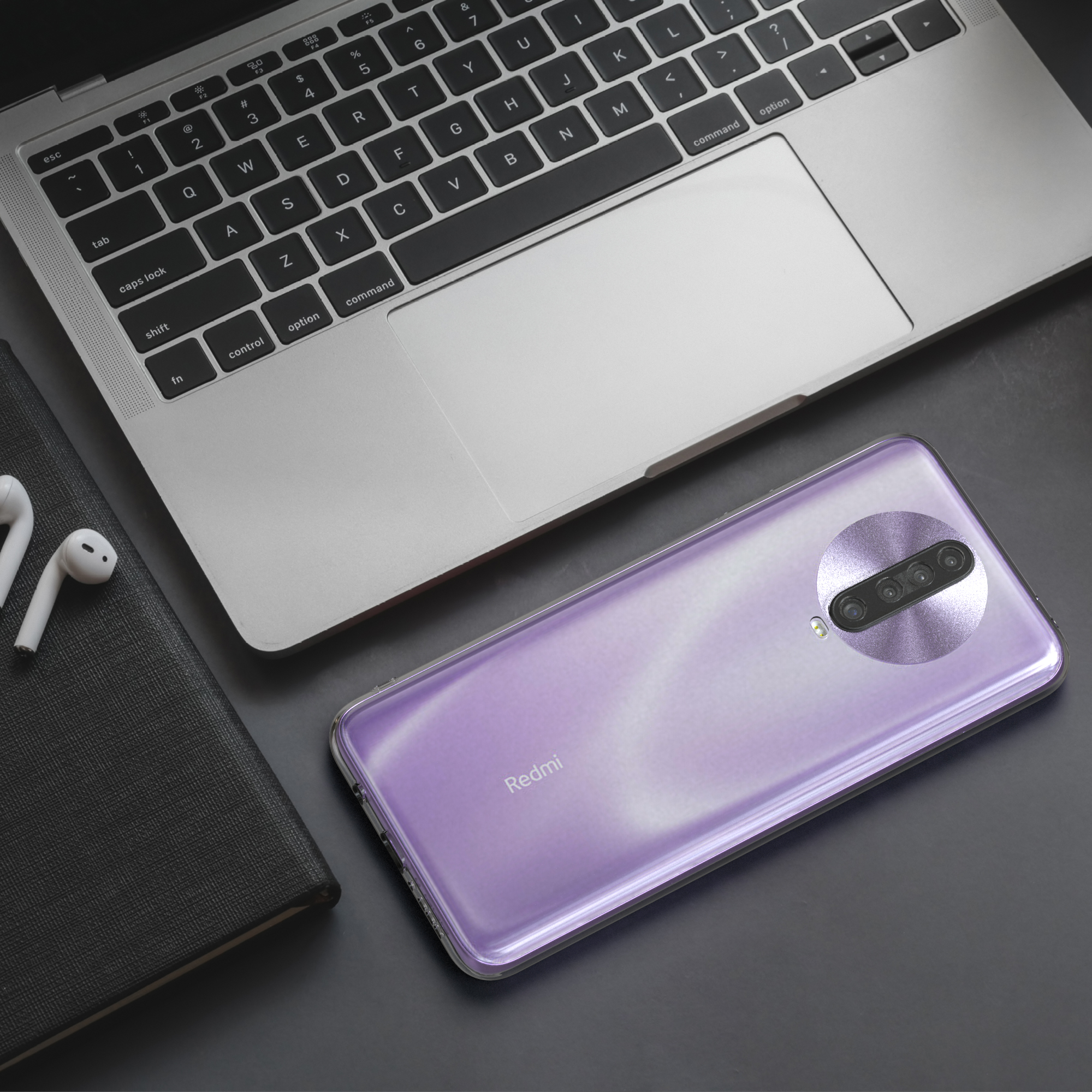 Redmi-Telefon auf einem schwarzen Schreibtisch neben einem Laptop, Notizbuch und weißen Ohrhörern. Das Telefon hat eine lilafarbene Rückseite.