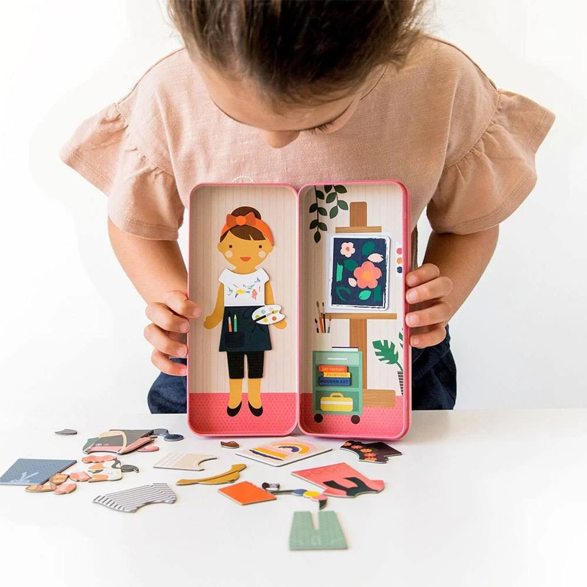 Ein Mädchen betrachtet ein Spielzeug mit einer Puppe und einem Miniatur-Kunstatelier. Verschiedene Spielzeugkleider sind verstreut.