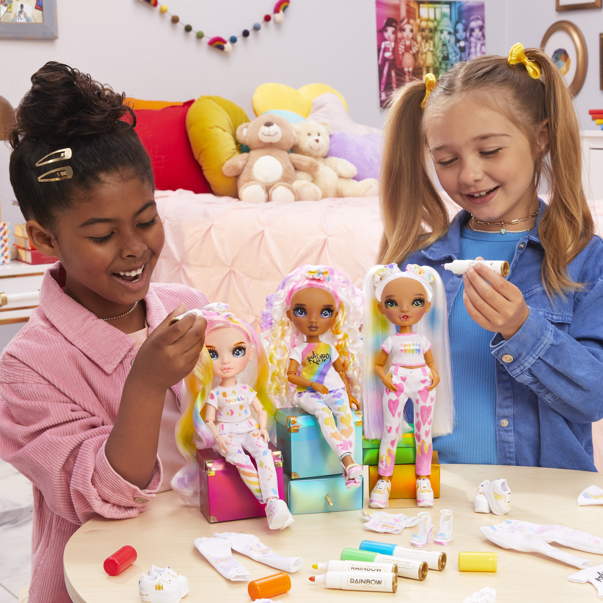 Dos chicas con muñecas Rainbow High. Están dibujando en la ropa de las muñecas. Hay tres muñecas y accesorios.