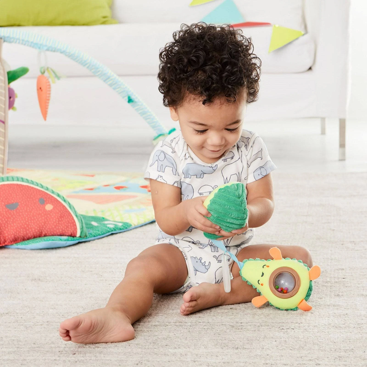 Ein Baby sitzt auf einem Teppich und hält ein grünes Avocado-Spielzeug. Das Baby trägt einen Strampler mit Elefantenmuster.