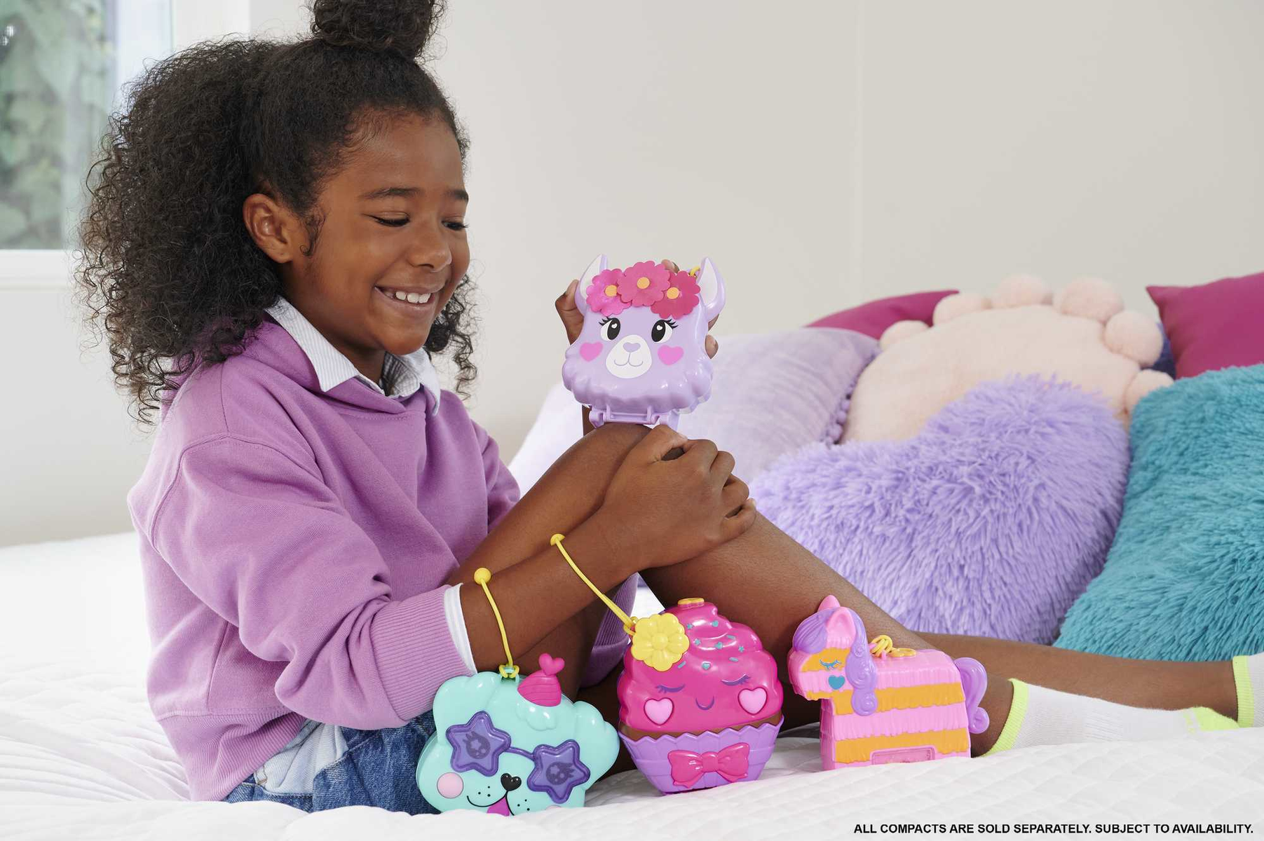 Ein Mädchen lächelt und hält drei Polly Pocket Spielzeuge auf einem Bett.