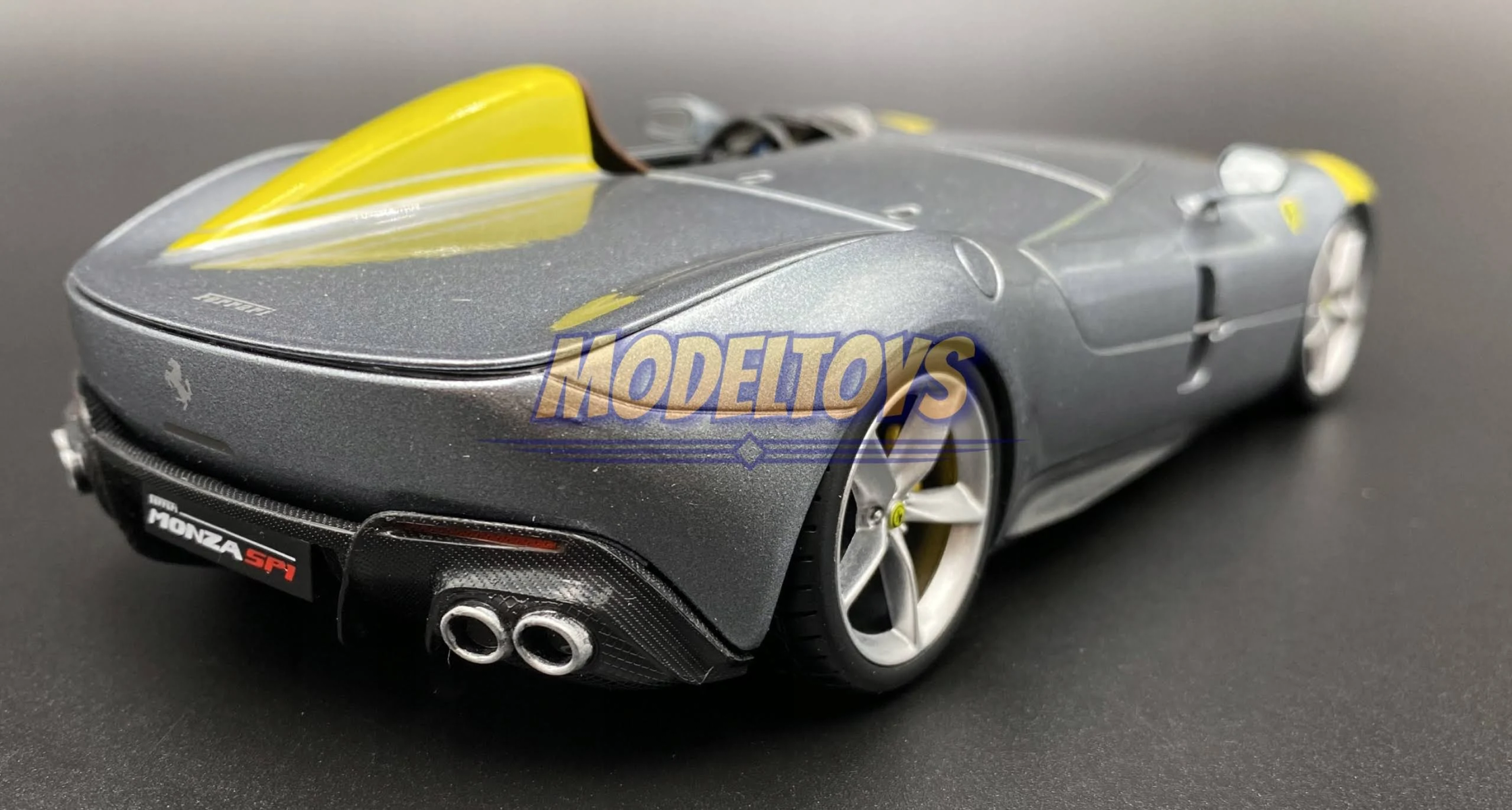 Un coche modelo Ferrari Monza SP1 gris y amarillo, vista trasera, sobre una superficie negra.