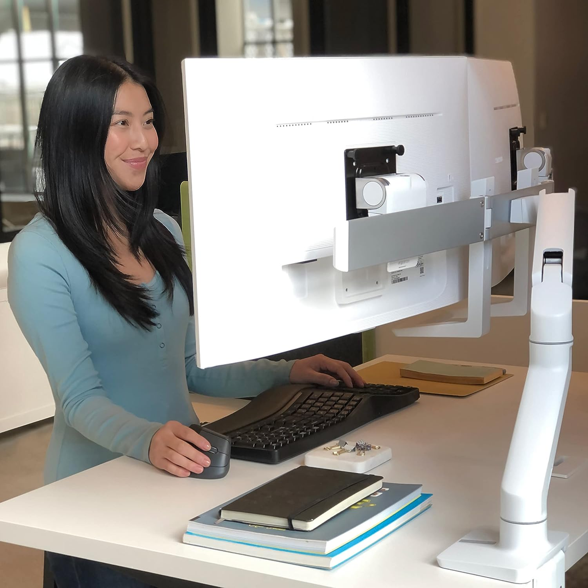 Woman using a computer with a large monitor, a keyboard, and a mouse, on a white desk. 