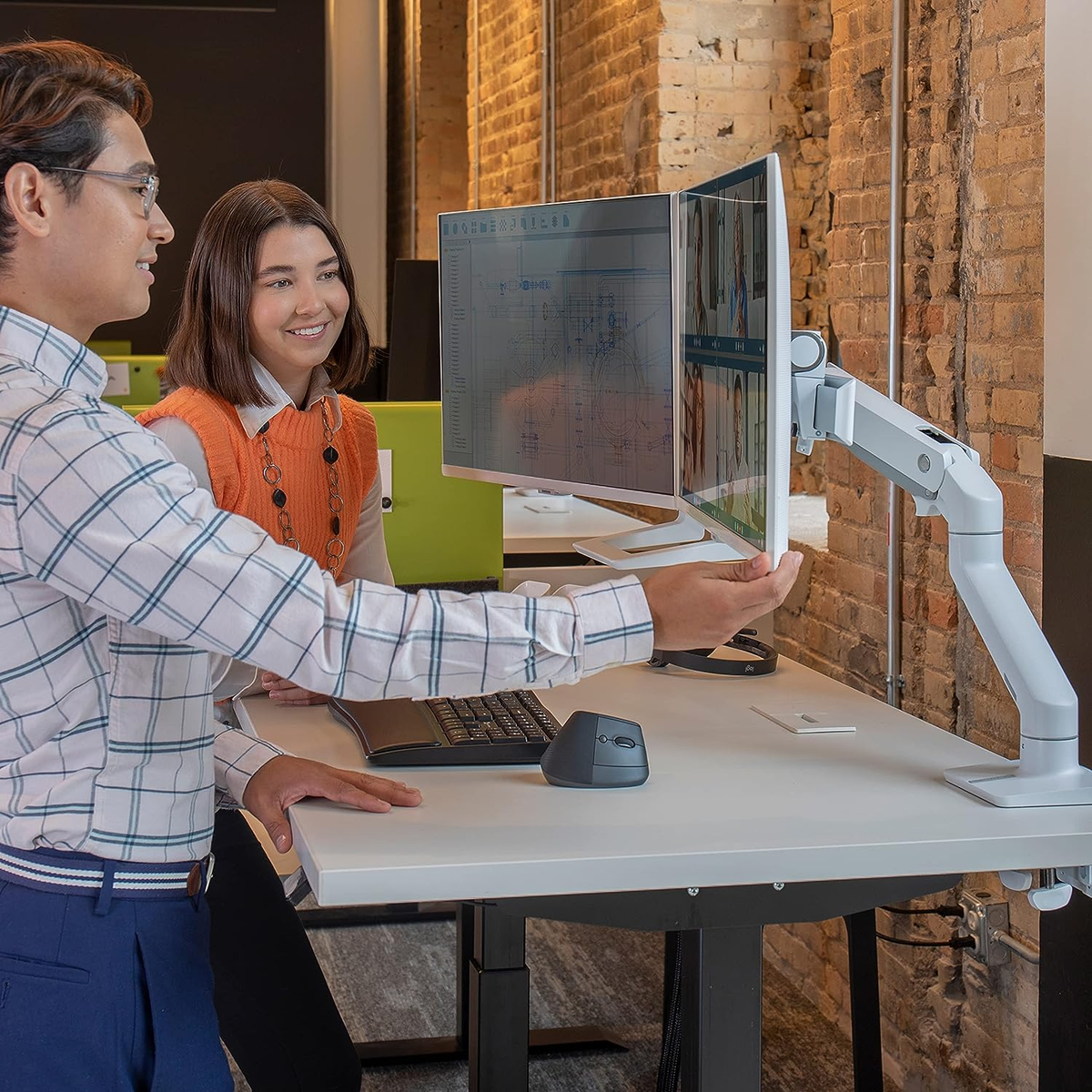 Two people at a desk looking at two computer monitors. Brick wall background. White desk.