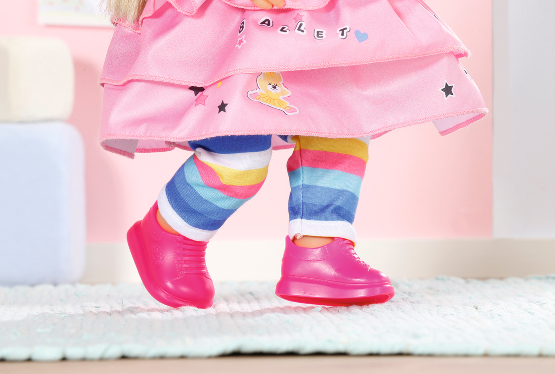 Puppenbeine und -füße in rosa Schuhen und bunten Leggings auf pinkfarbenem Hintergrund.