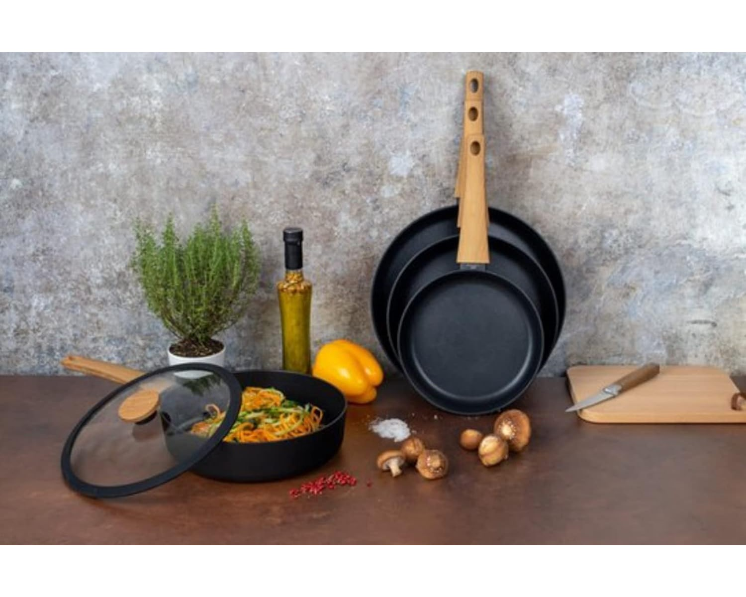 Schwarze Pfannen mit Holzgriffen, Gemüse, Ölflasche und Pilze auf einem braunen Tisch.