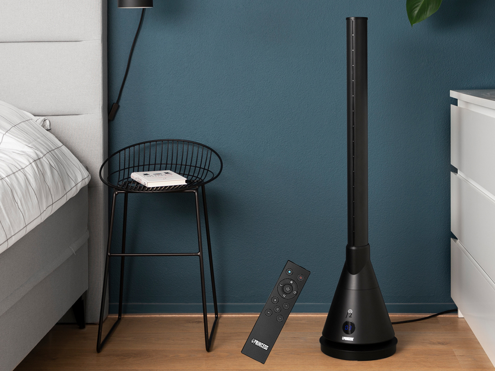 Un ventilatore a torre nero e un telecomando. Un piccolo sgabello e un mobile bianco sono sullo sfondo.