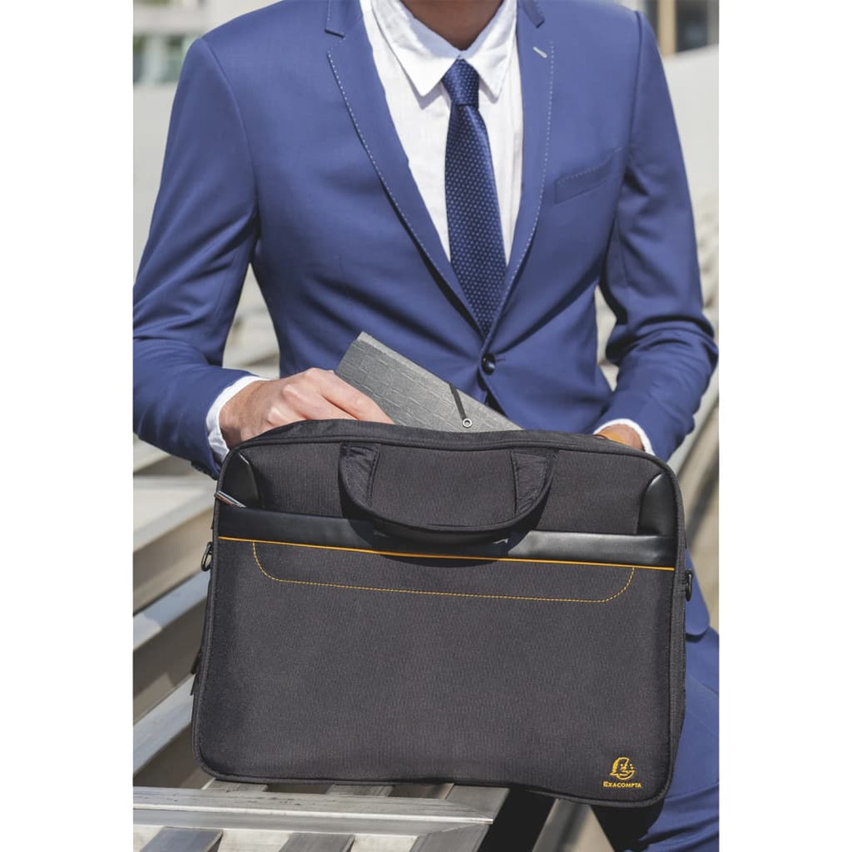 Una persona con traje azul sostiene un maletín negro. El maletín tiene un asa y costuras amarillas.