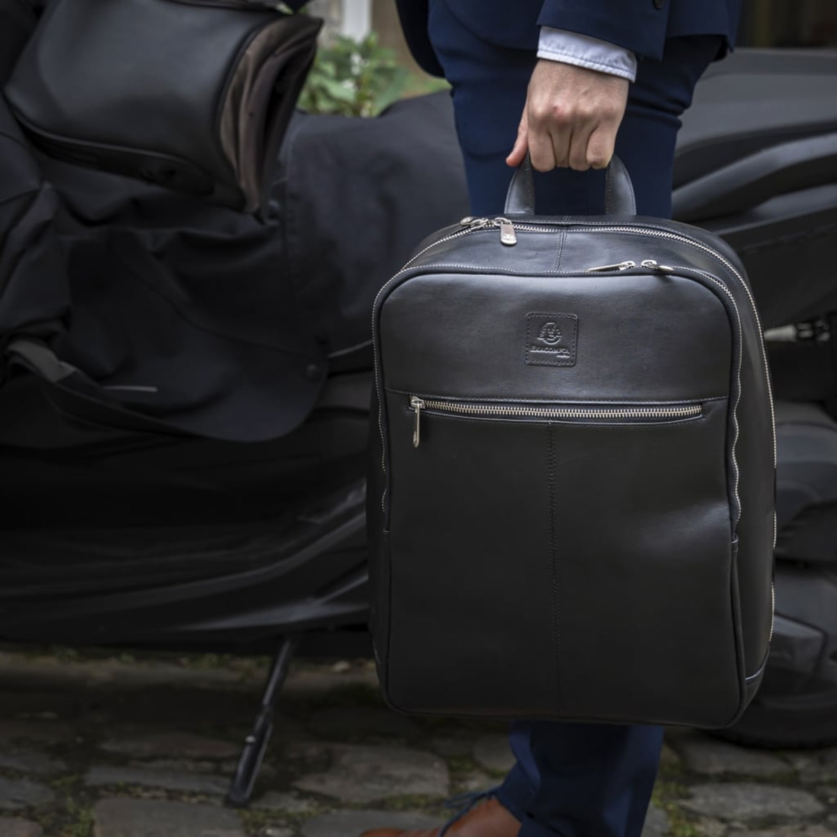 Un hombre en traje sostiene una mochila de cuero negro con cremallera y asa.