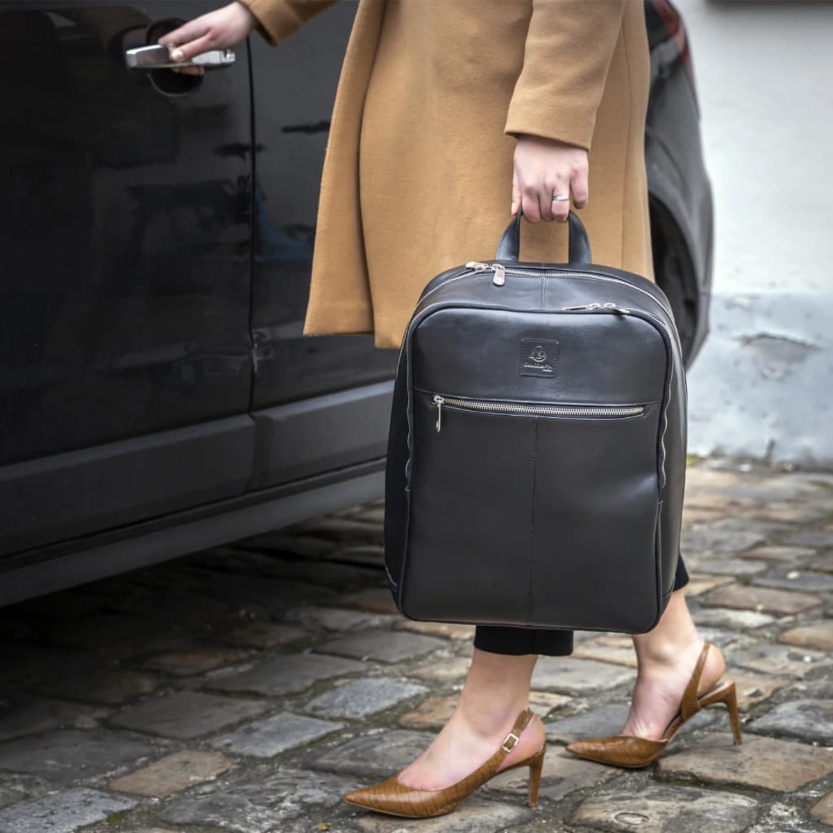 Una persona con abrigo beige y tacones, llevando una mochila negra, junto a un coche negro.