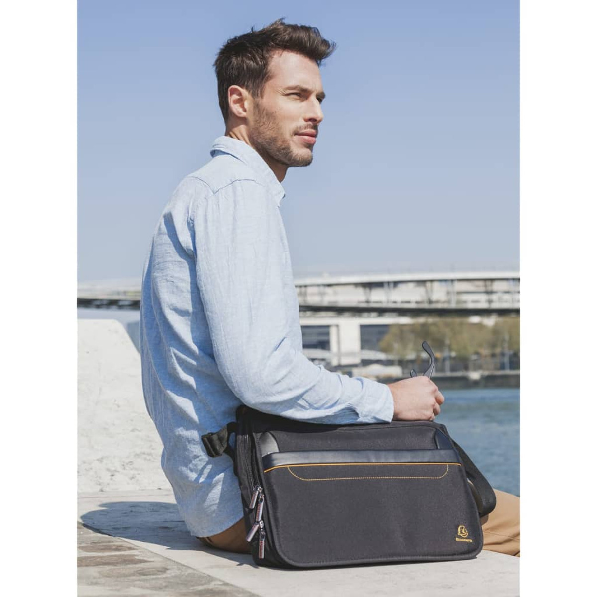 Hombre con bolso, mirando hacia un lado. Lleva una camisa azul. Un río y un puente al fondo.