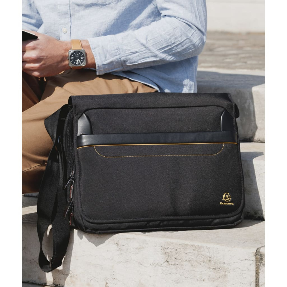 Hombre sentado con bolso mensajero negro con raya dorada, sosteniendo un reloj.