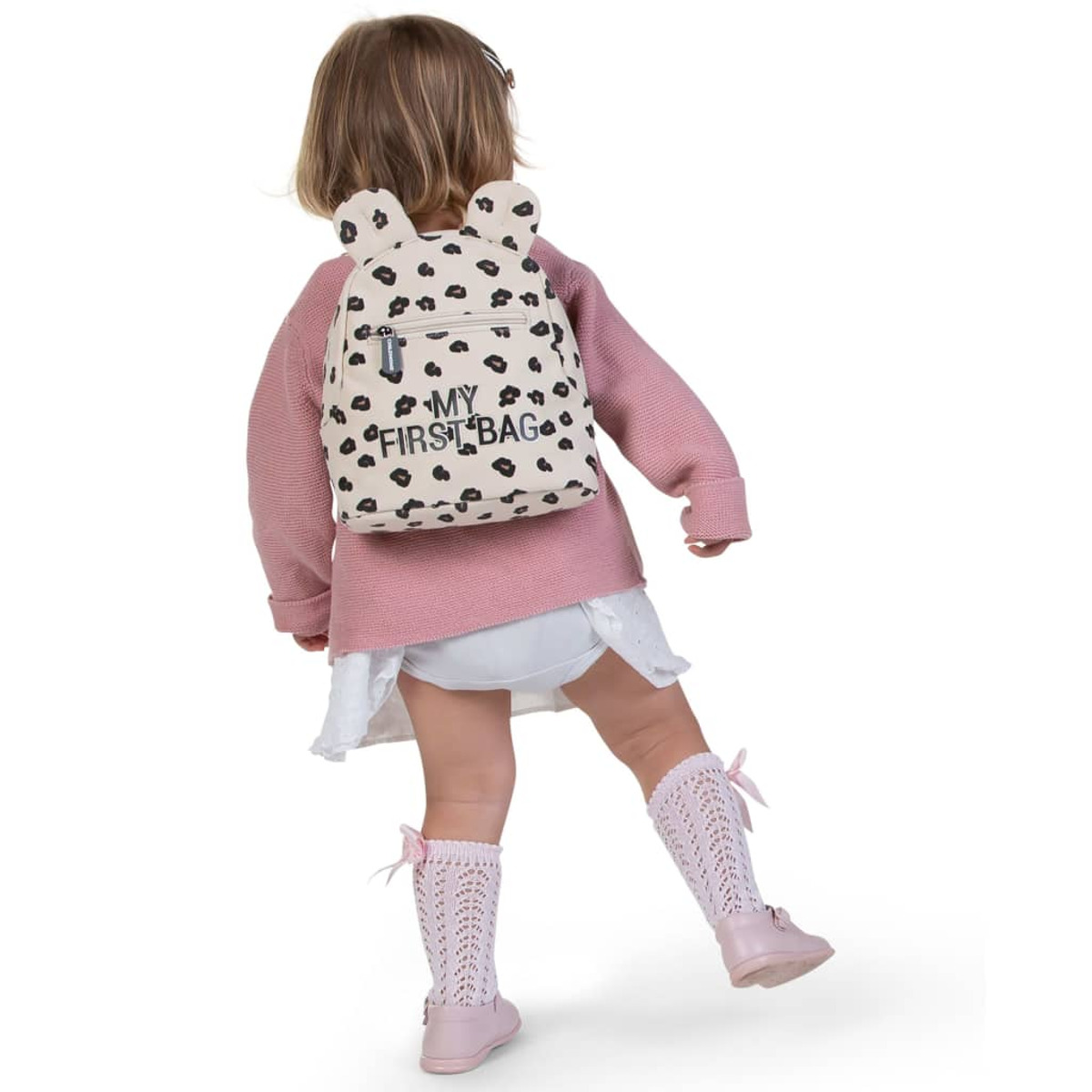 Ein junges Mädchen in rosa Pullover und weißem Kleid mit einem Rucksack mit der Aufschrift 'My First Bag'.