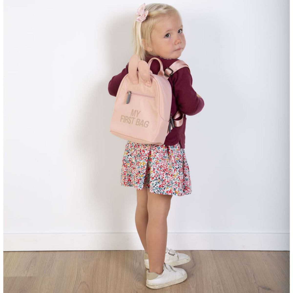 Ein junges Mädchen in einem weinroten Pullover und einem Blumenrock mit einem rosa Rucksack.