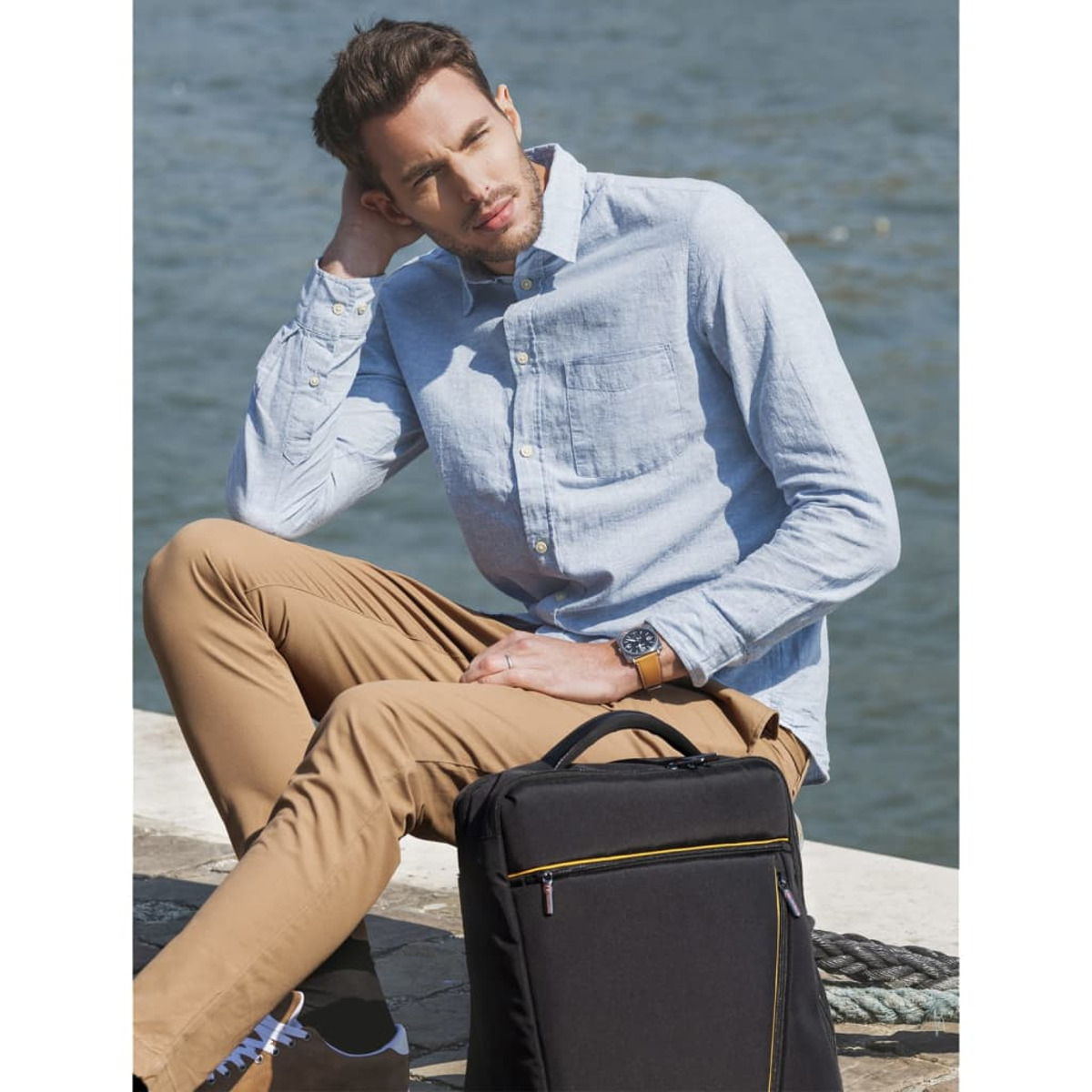 Hombre sentado. Camisa celeste, pantalón marrón, bolso negro y reloj. Río y fondo borroso.