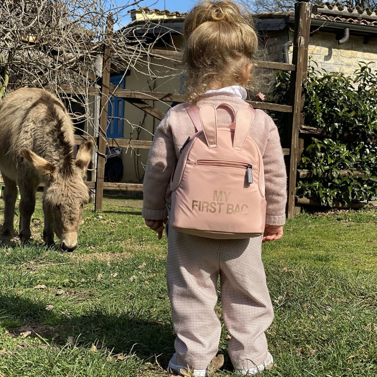 Ein junges Mädchen mit einem rosa Rucksack steht in einem grasbewachsenen Garten. Ein Esel im Hintergrund.