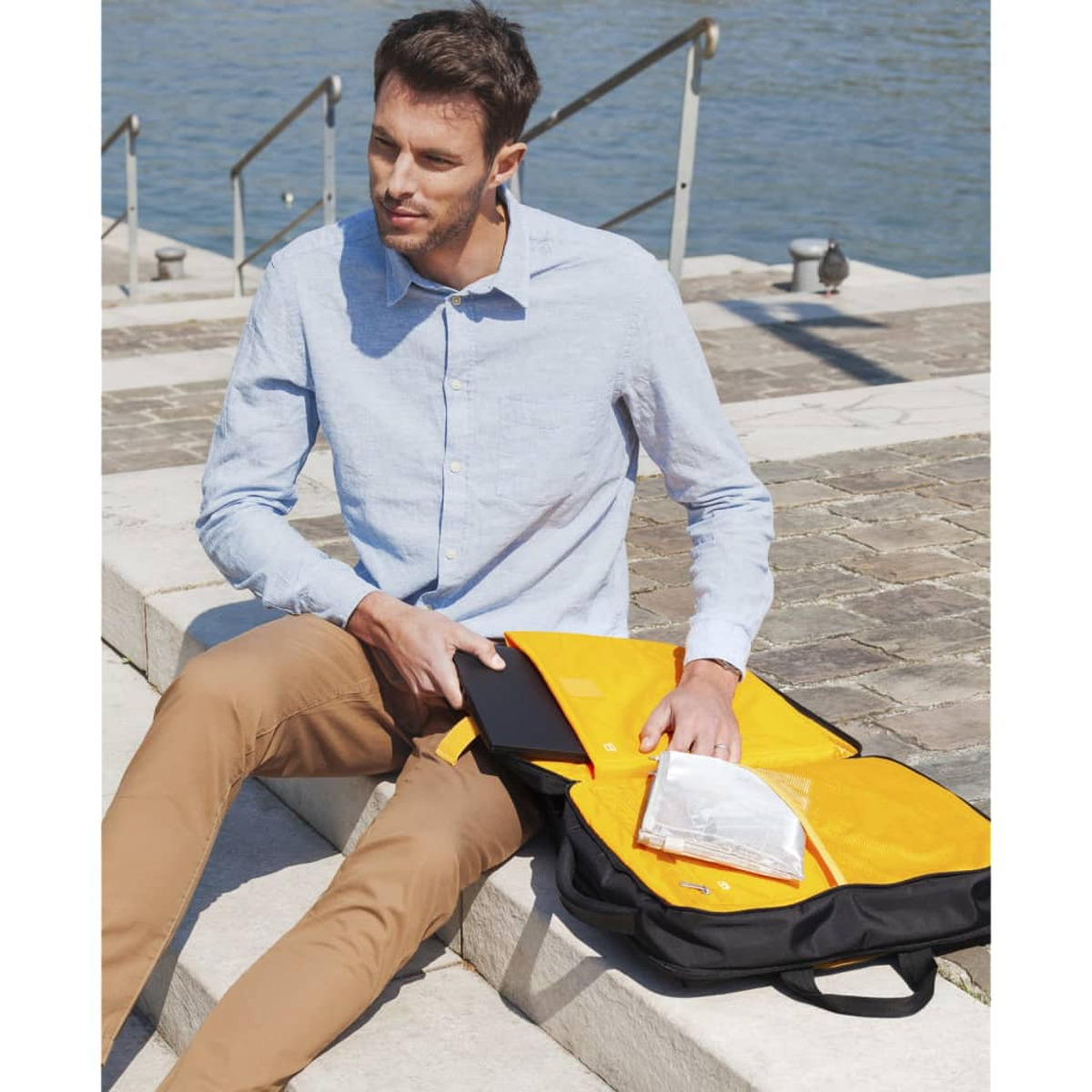 Hombre con camisa azul y pantalones marrones sentado en escalones. Bolso negro con objeto blanco.