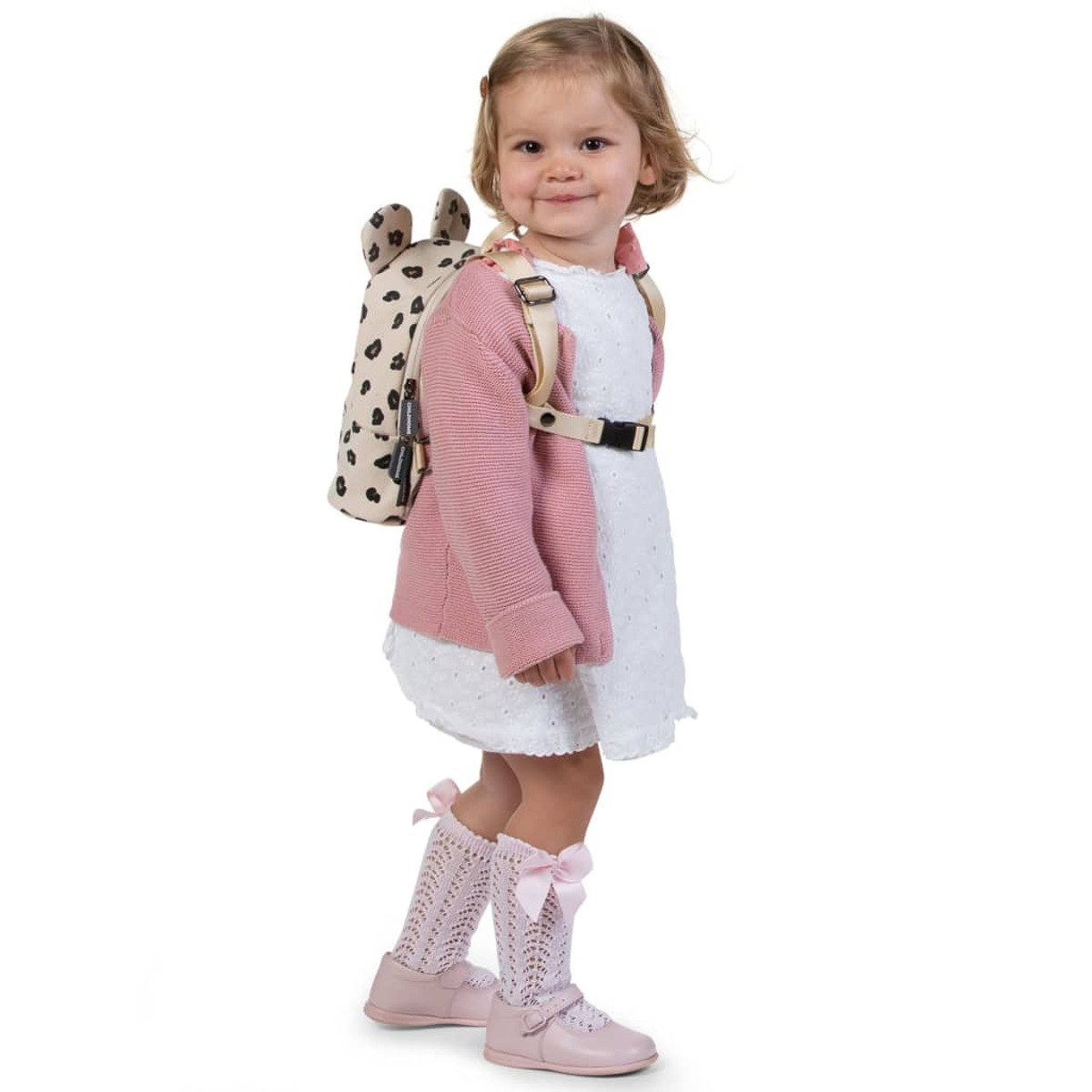 Ein junges Mädchen mit Rucksack, rosa Pullover und weißem Kleid, lächelnd.