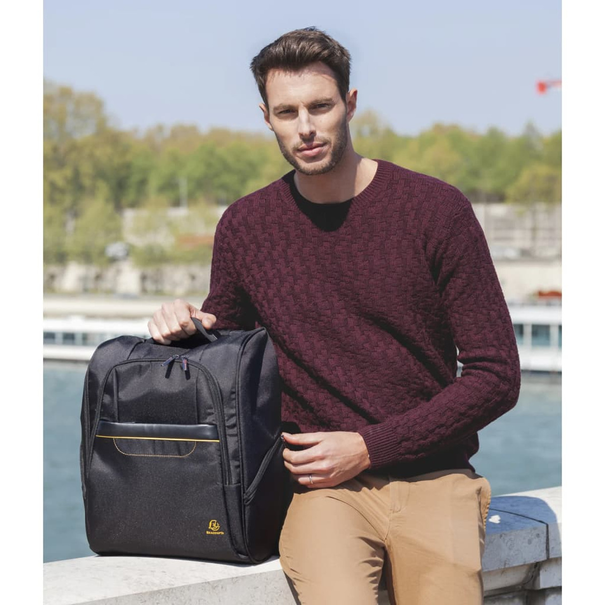 Hombre con suéter burdeos y pantalones beige, sosteniendo una mochila negra junto al agua.