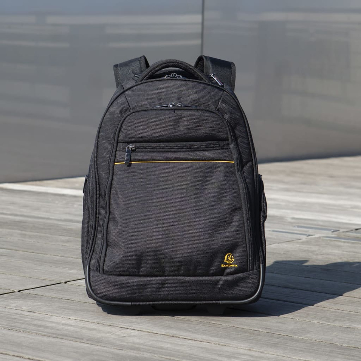 Una mochila negra con cremallera amarilla. Se coloca en una cubierta de madera, con un fondo gris.