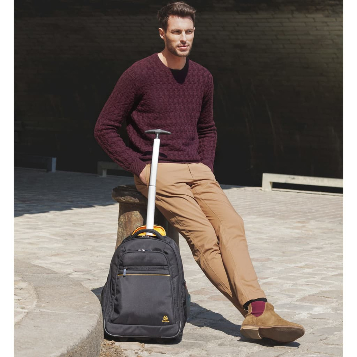 Hombre con mochila apoyado en estructura de piedra, con suéter y pantalones color caqui.