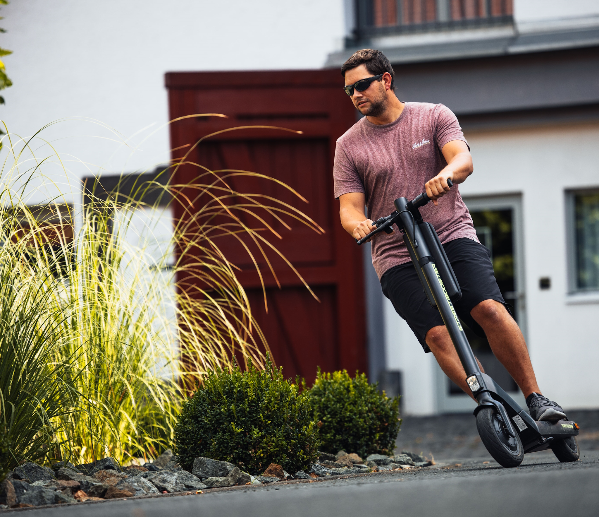 Ein Mann mit Sonnenbrille fährt mit einem Elektroroller auf einer asphaltierten Straße, in der Nähe von Büschen.