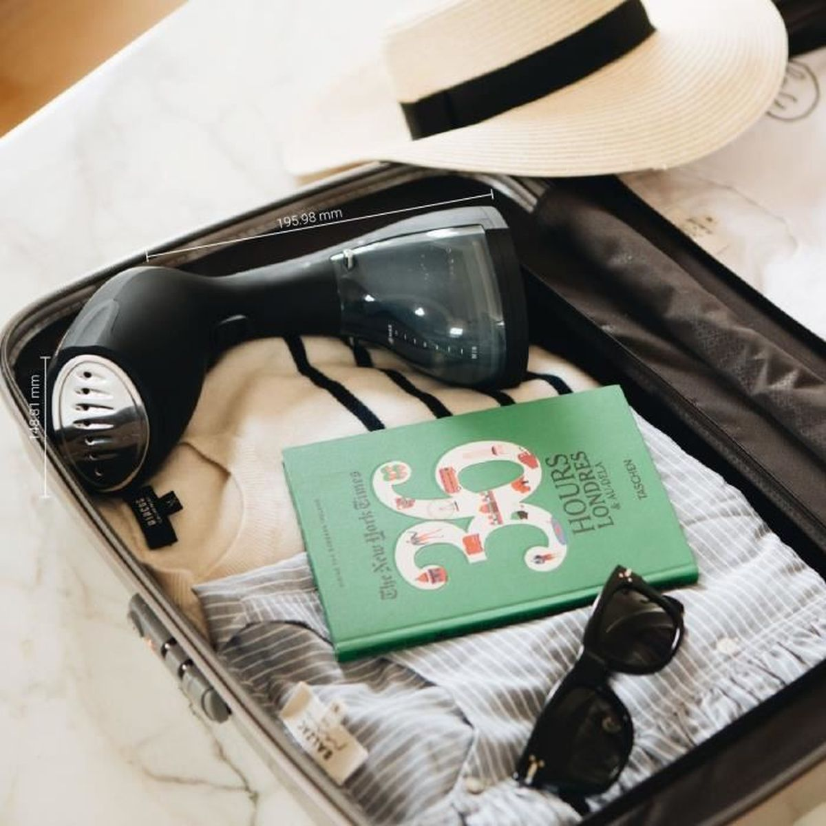 Valise ouverte avec vêtements, livre, lunettes de soleil et défroisseur. Un chapeau est dessus.