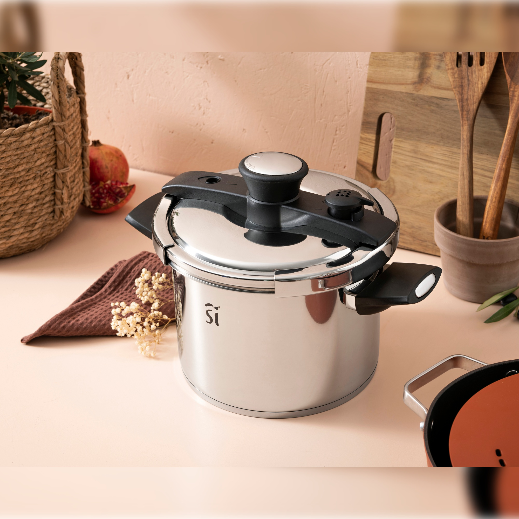 Una olla a presión de acero inoxidable sobre una superficie rosa, con utensilios de madera al fondo.