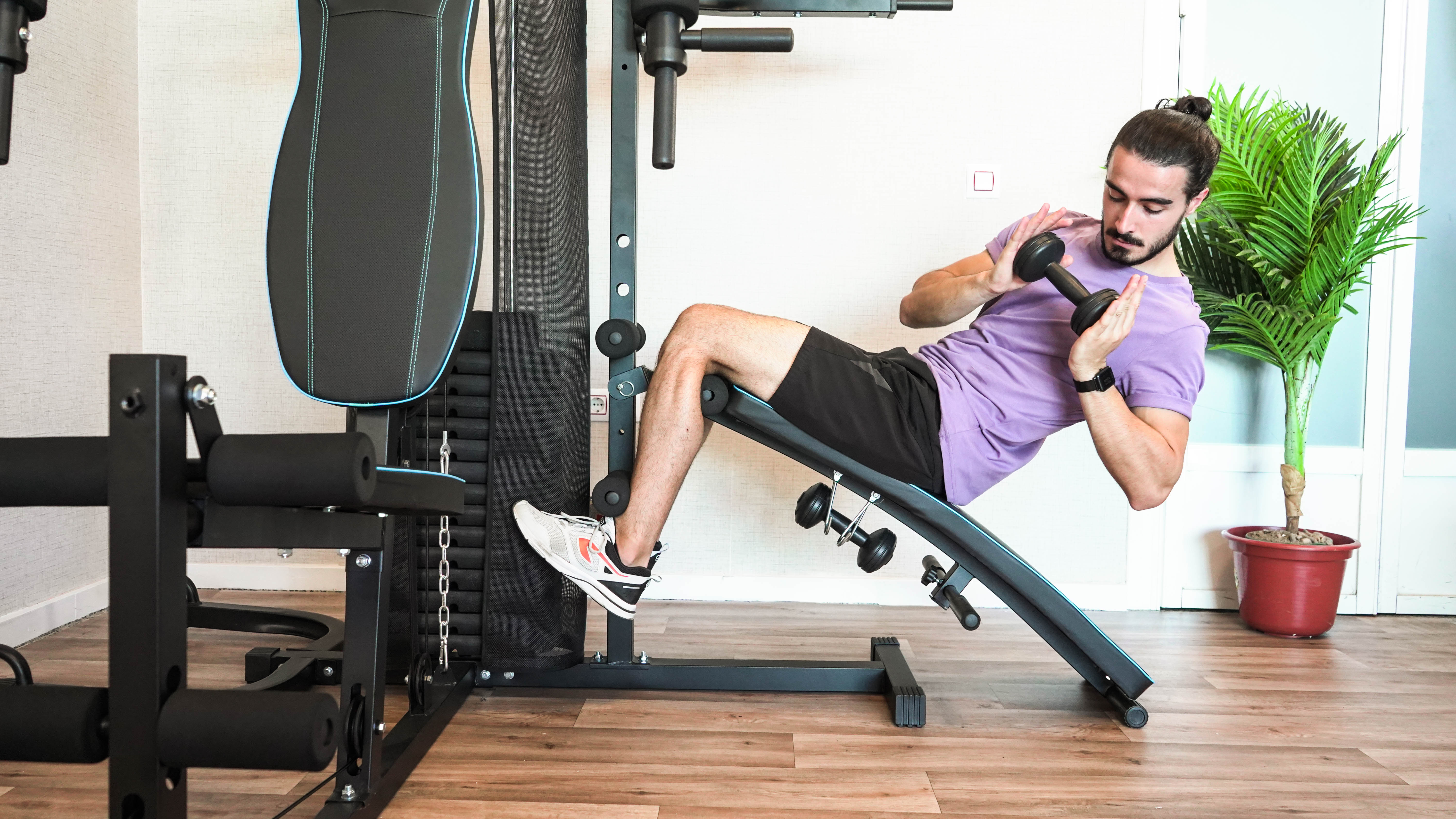 Hombre haciendo ejercicio con una mancuerna en un banco inclinado. Equipo de gimnasio visible. Ambiente de la habitación.