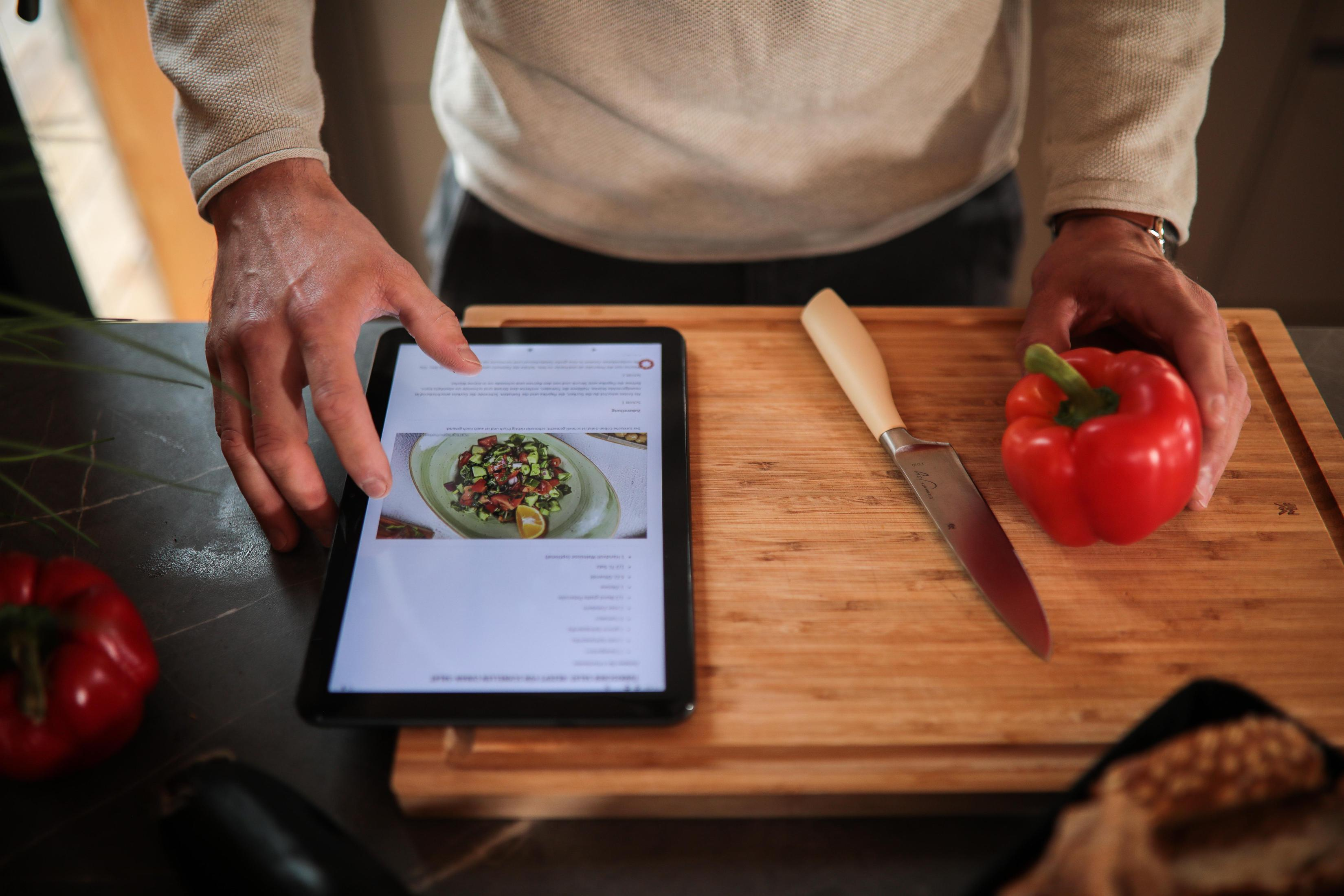 Eine Person benutzt ein Tablet mit einem Rezept, mit Essen und einem Messer auf einem Brett.