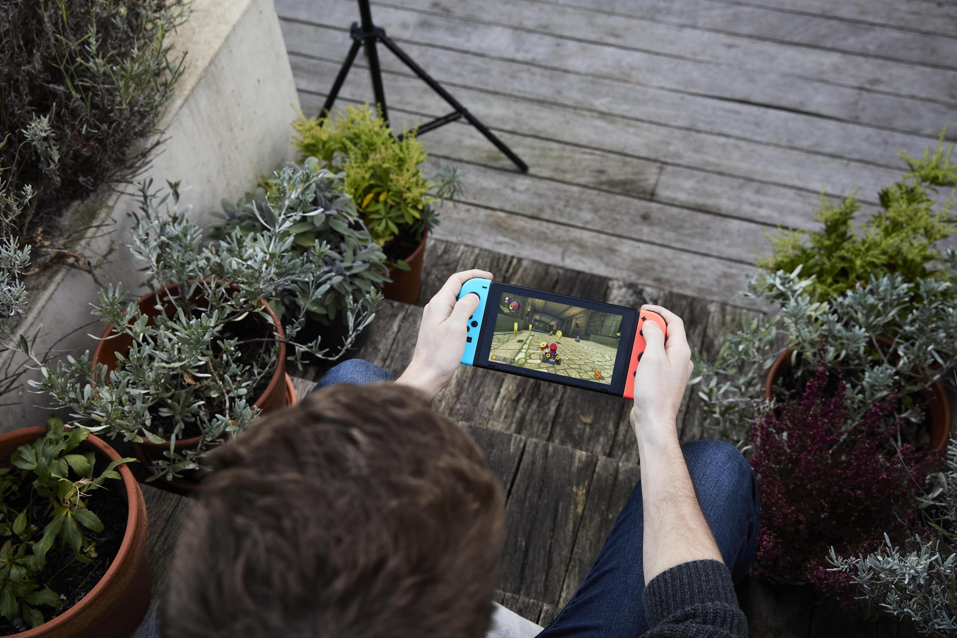 Person spielt Nintendo Switch im Freien. Handheld-Konsole mit blauen und roten Controllern. Holzdeck.