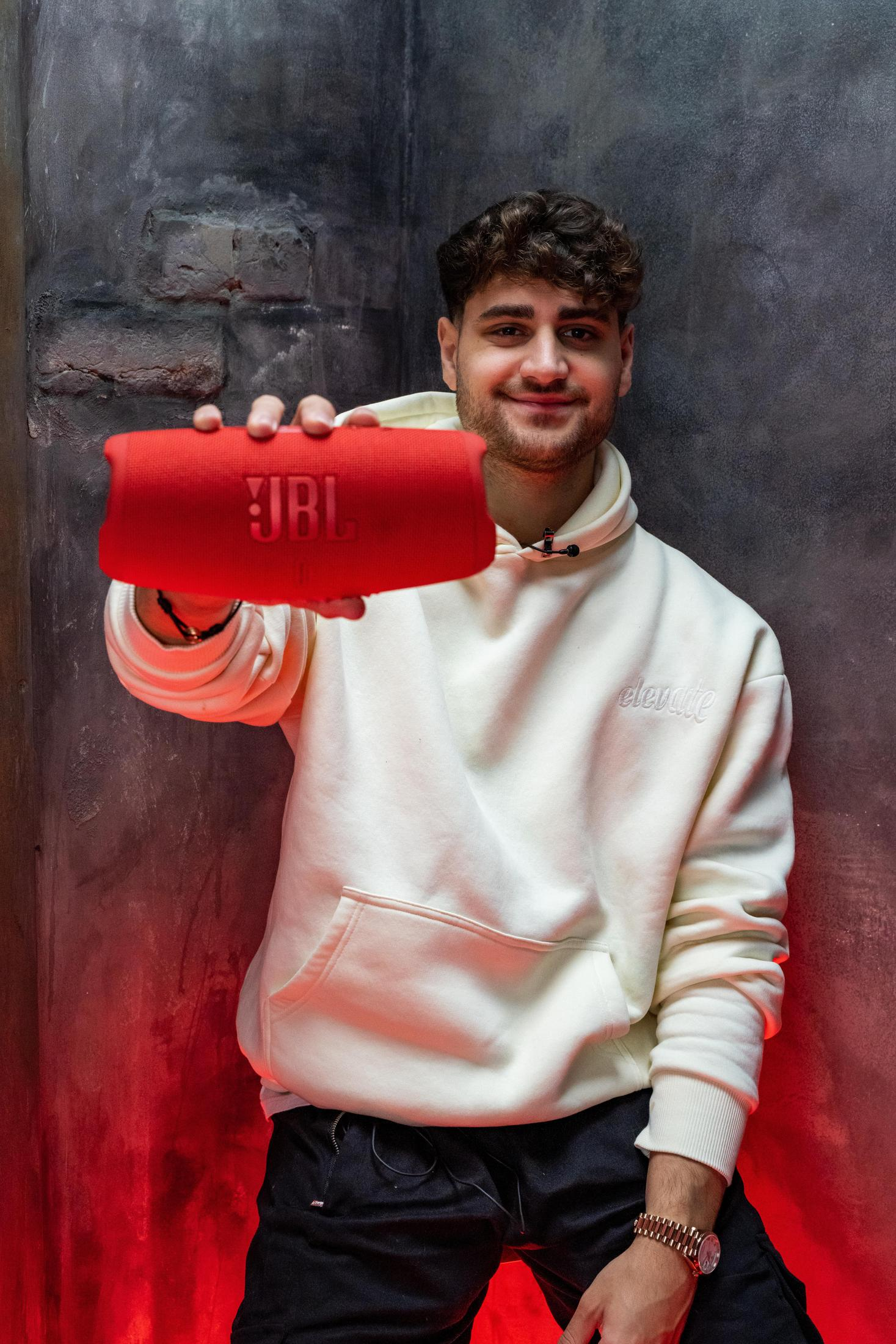 Mężczyzna trzymający czerwony głośnik JBL, w białej bluzie z kapturem, uśmiecha się do kamery.