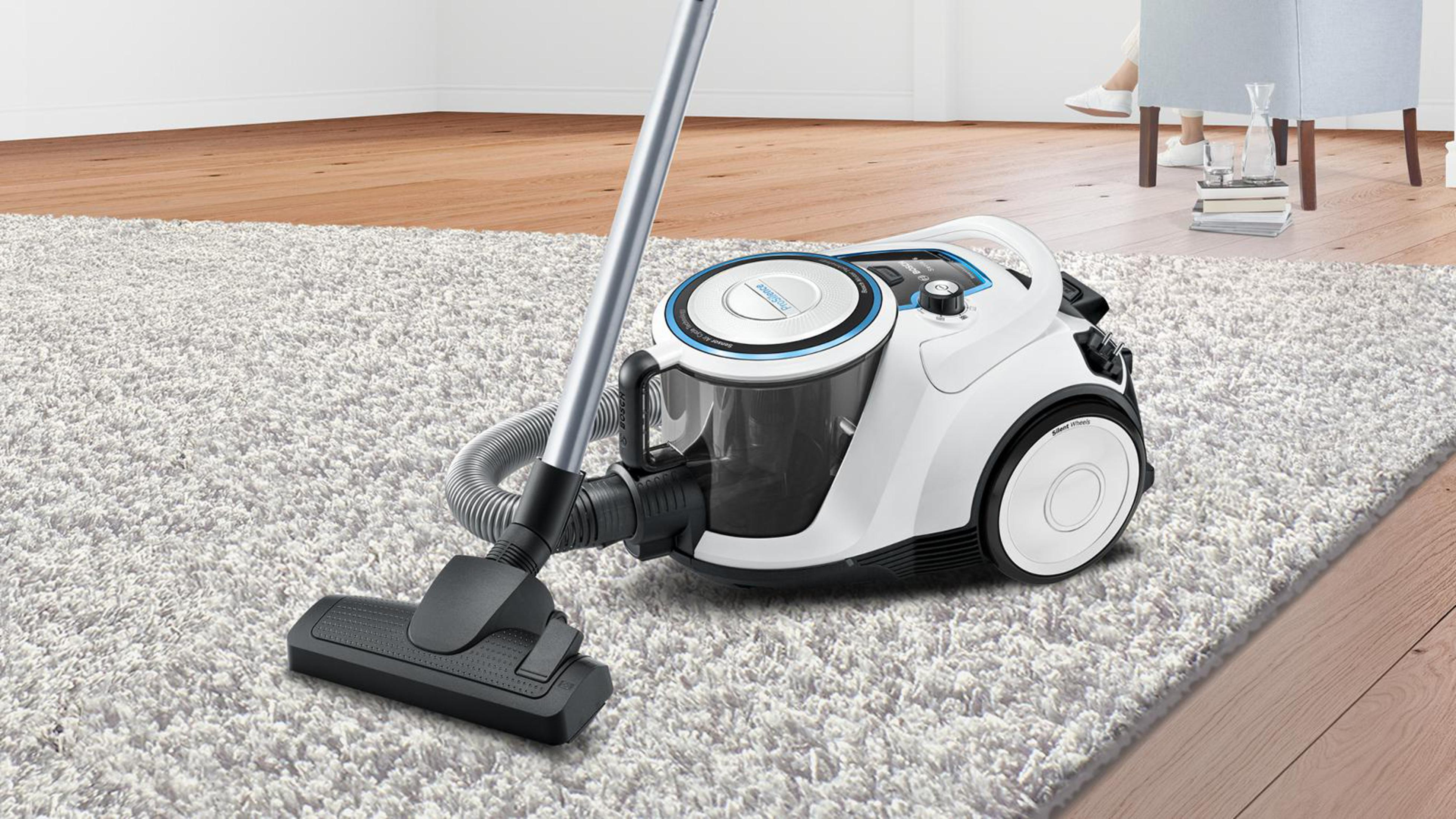 Odkurzacz na białym dywanie z drewnianą podłogą i osobą w tle.